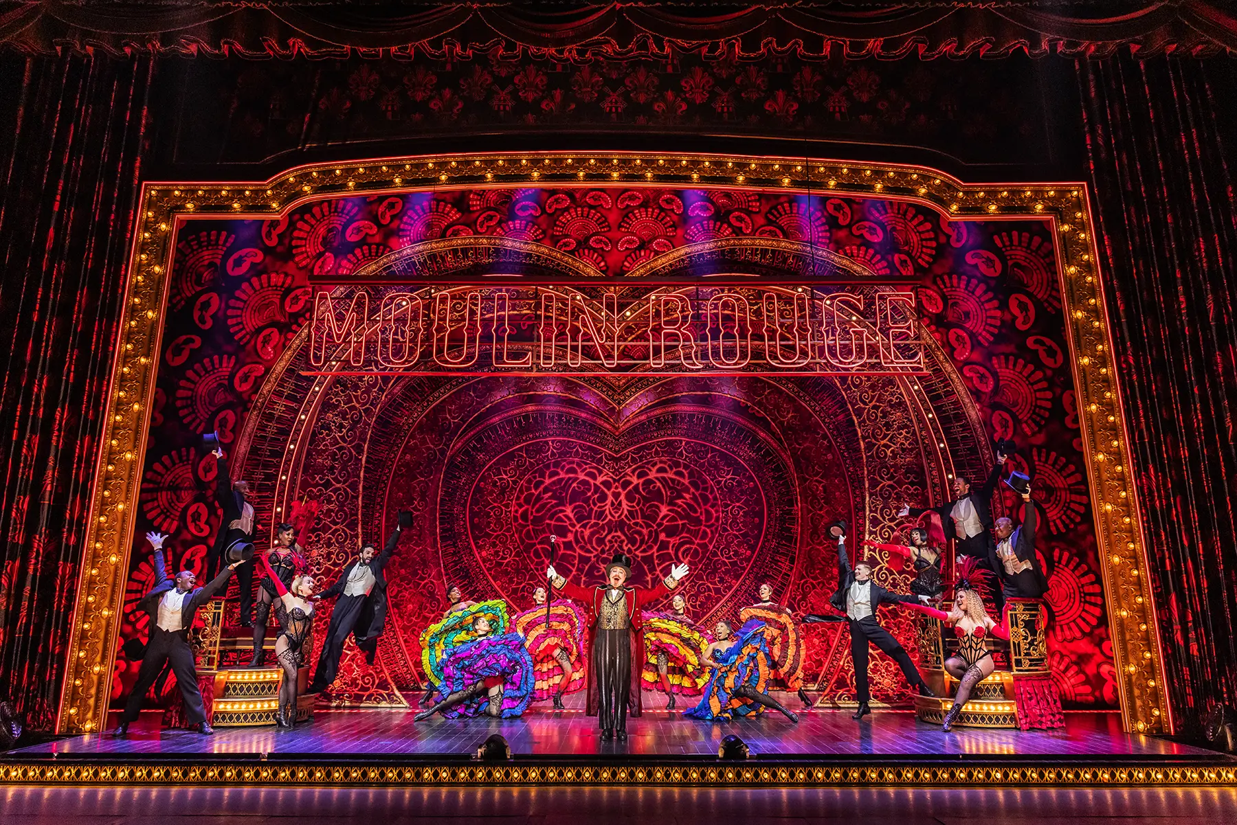 Scene from Moulin Rouge! The Musical's North American Tour, with dancers in colorful costumes performing on an ornate stage featuring a neon “MOULIN ROUGE” sign and heart-shaped backdrop.