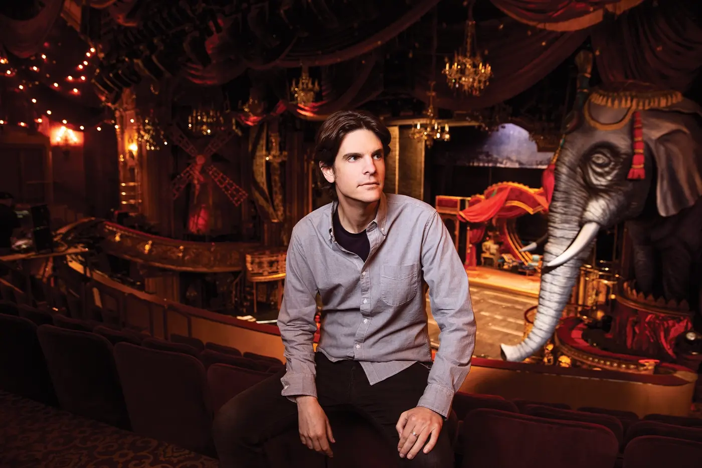 Alex Timbers, director of Moulin Rouge! The Musical sitting in front of the set of the production, with a giant blue elephant visible on the right.