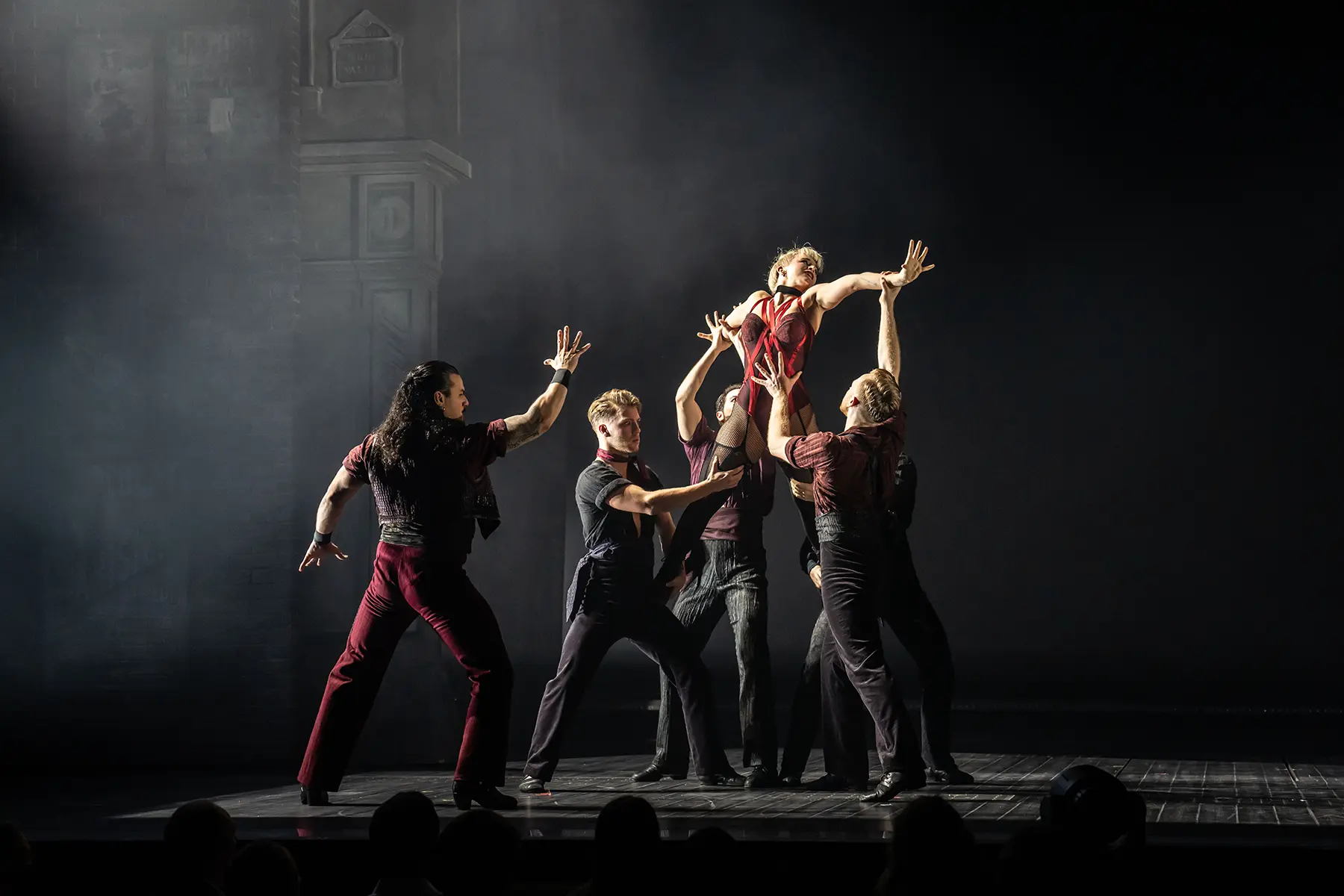A dramatic moment from 'El Tango de Roxanne' in Moulin Rouge! The Musical (Germany). A performer in a striking red costume is lifted into the air by several ensemble members. The stage is dimly lit, with dark tones and architectural elements visible on the left, enhancing the intensity and passion of the scene.