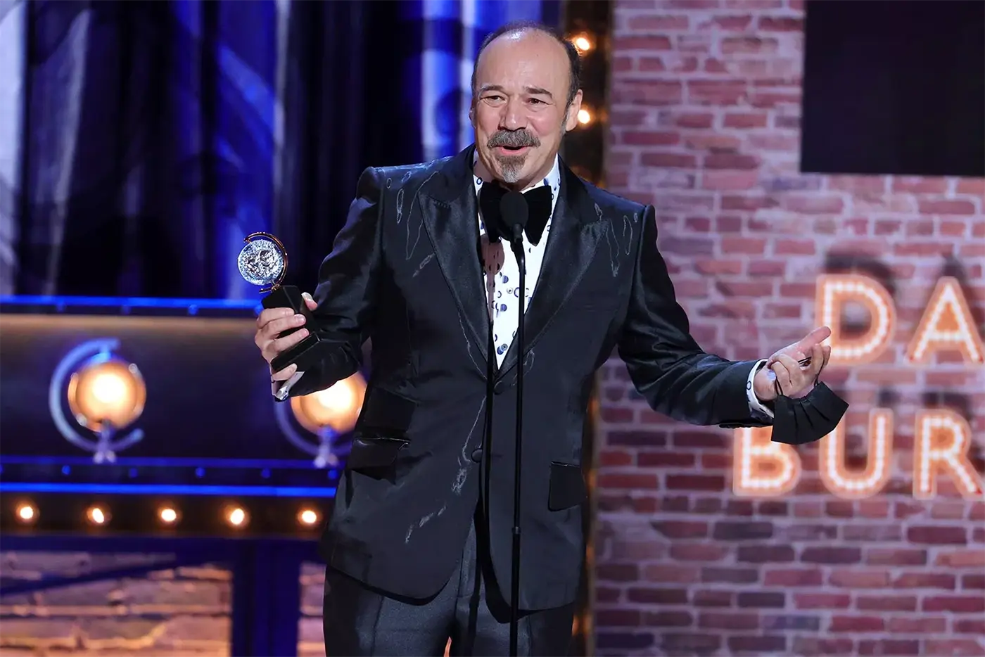 Danny Burstein on stage accepting his Tony Award for Moulin Rouge! The Musical against a backdrop of blue curtains and stage lights.