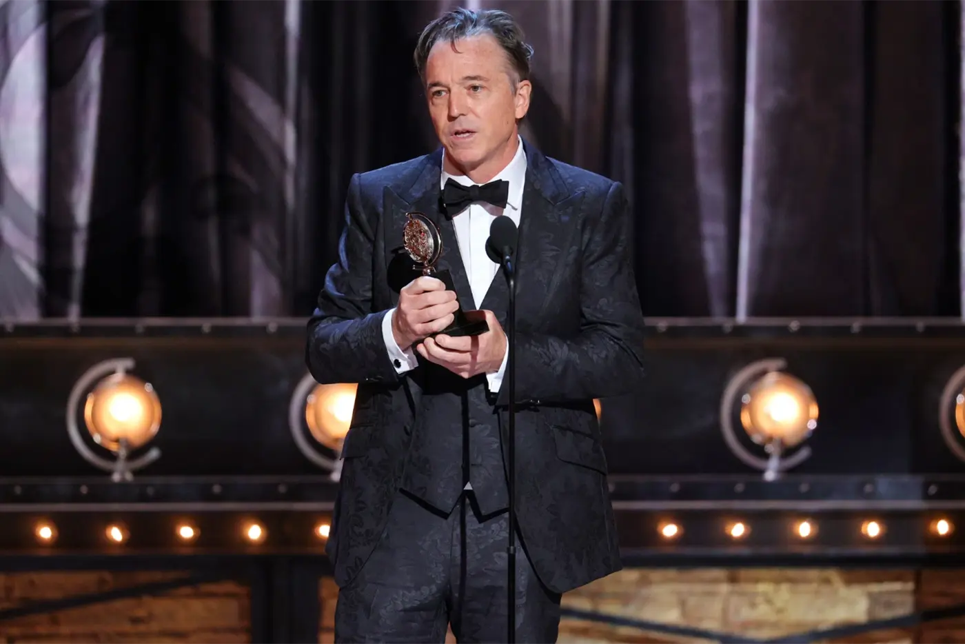 Derek McLane on stage accepting his Tony Award for Moulin Rouge! The Musical against a backdrop of blue curtains and stage lights.