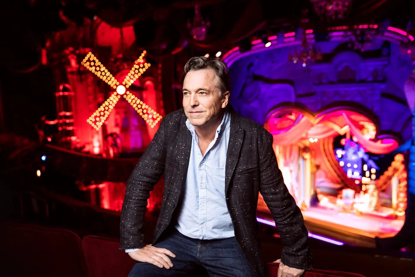 Derek McLane, scenic designer of Moulin Rouge! The Musical sitting in front of the lavish set, illuminated pink and purple.