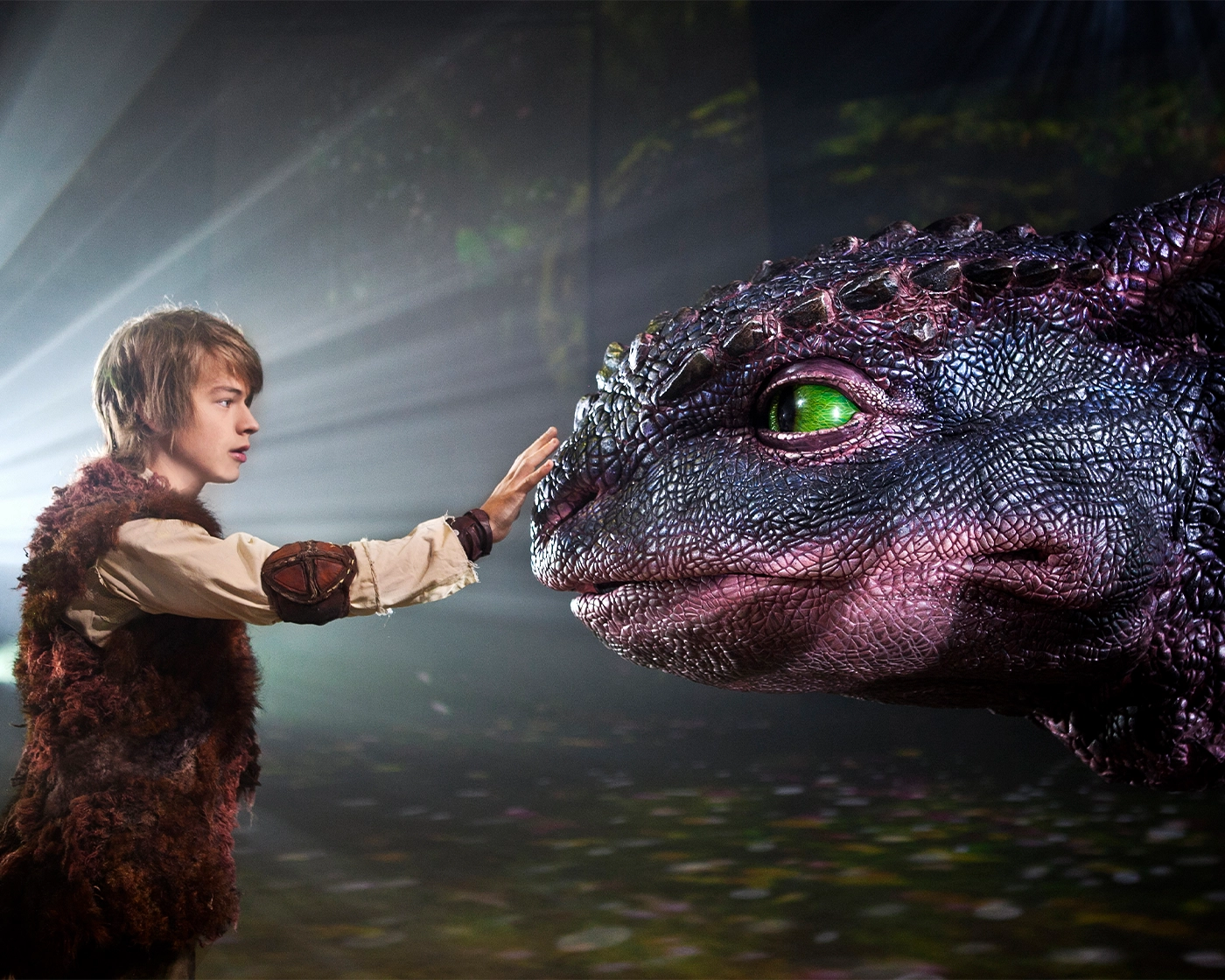 A young actor dressed in a fur vest and medieval-style costume gently reaching out to touch the nose of a large, lifelike dragon puppet with green eyes, under dramatic stage lighting.