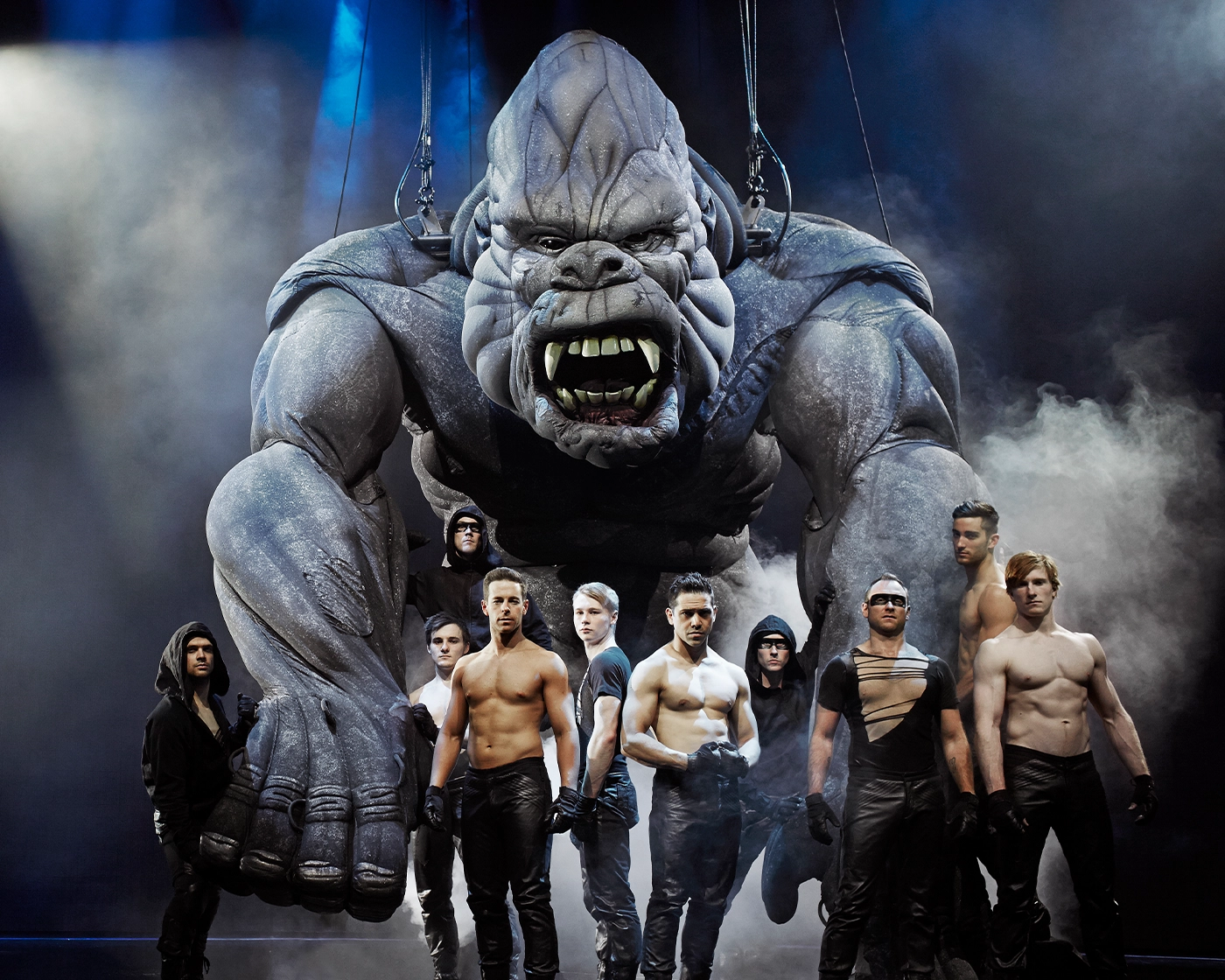 Scene from the Australian production of King Kong, featuring a large suspended puppet surrounded by performers under dramatic lighting and fog effects.