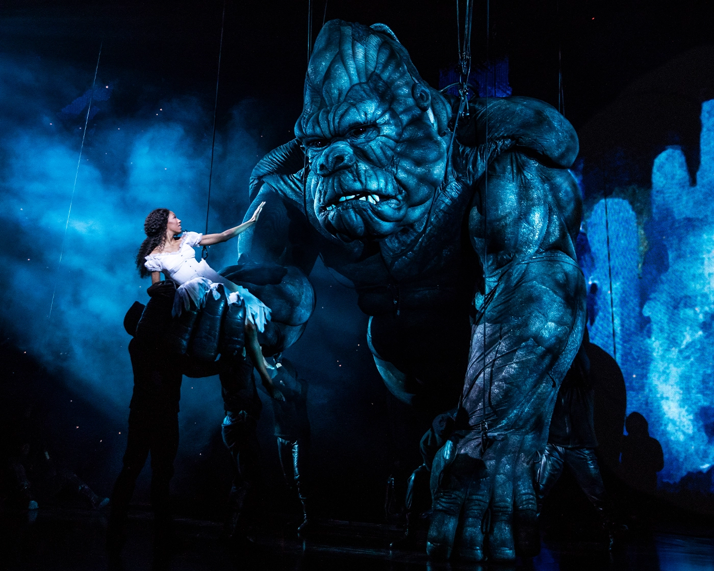 Scene from the Broadway production of King Kong, featuring the giant puppet holding a woman in white, with dramatic blue lighting and visible puppeteers.