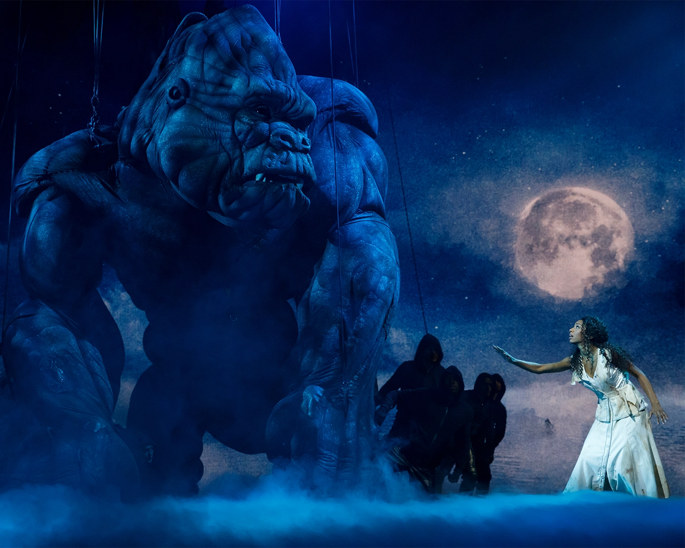 Scene from the Broadway production of King Kong, featuring a blue-lit gorilla puppet reaching toward a person in a white dress, set against a night sky with a full moon.