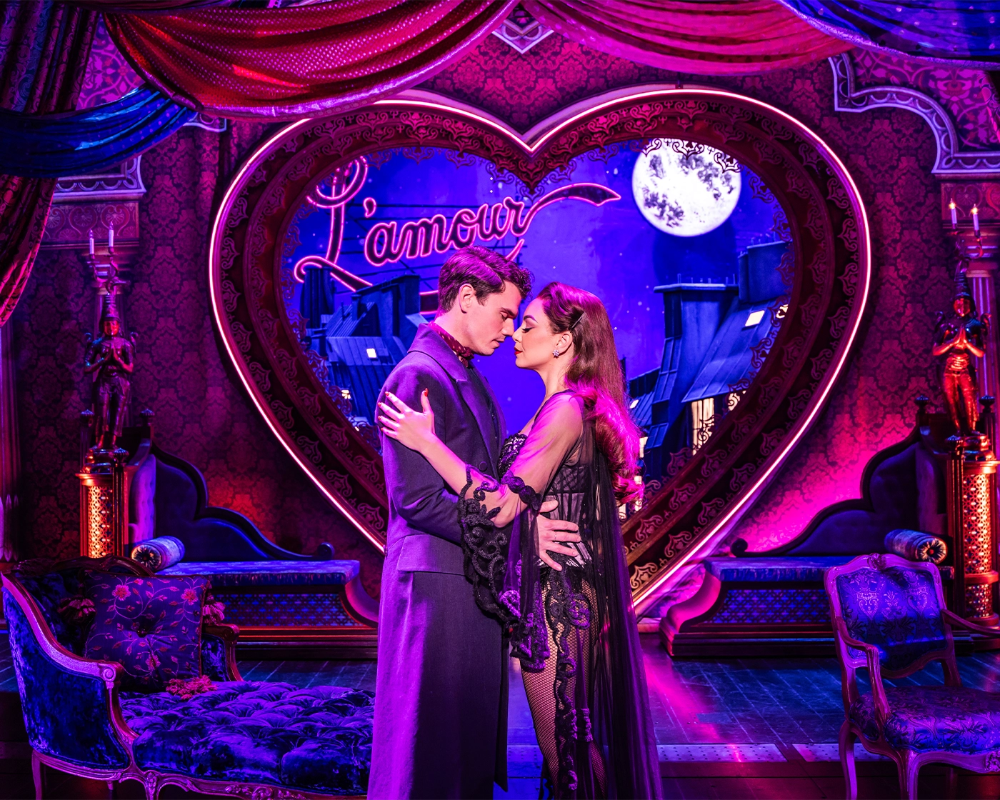 Scene from the Moulin Rouge North American tour in Satine’s dressing room, with two characters embracing in front of a heart-shaped “L’amour” sign, surrounded by opulent furnishings and purple drapes.