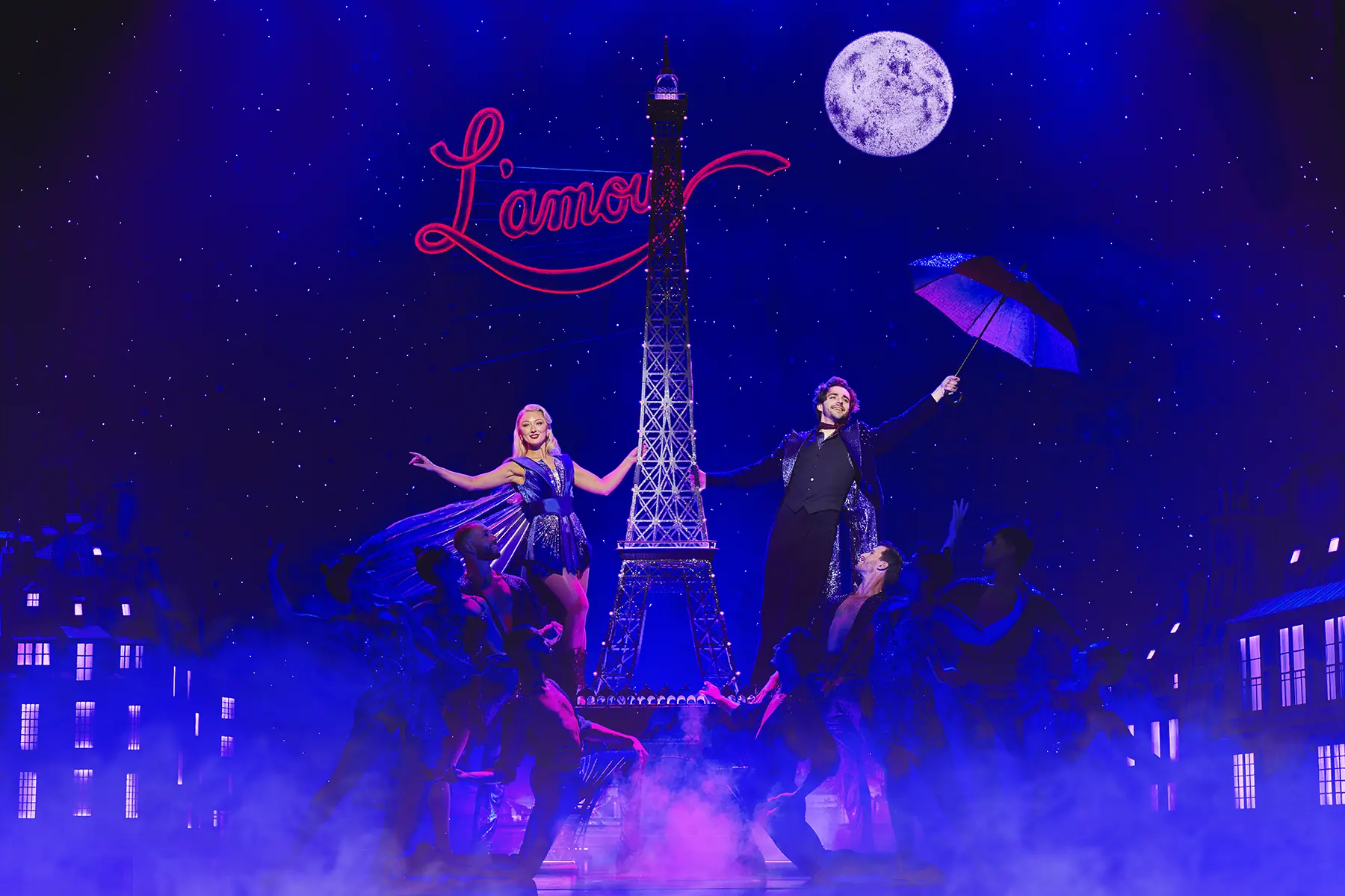 Scene from Moulin Rouge! The Musical World Tour during “Elephant Love Medley,” with two main characters beside a central Eiffel Tower structure, one holding a sparkly umbrella, under starry lights and a glowing “L’amour” sign.