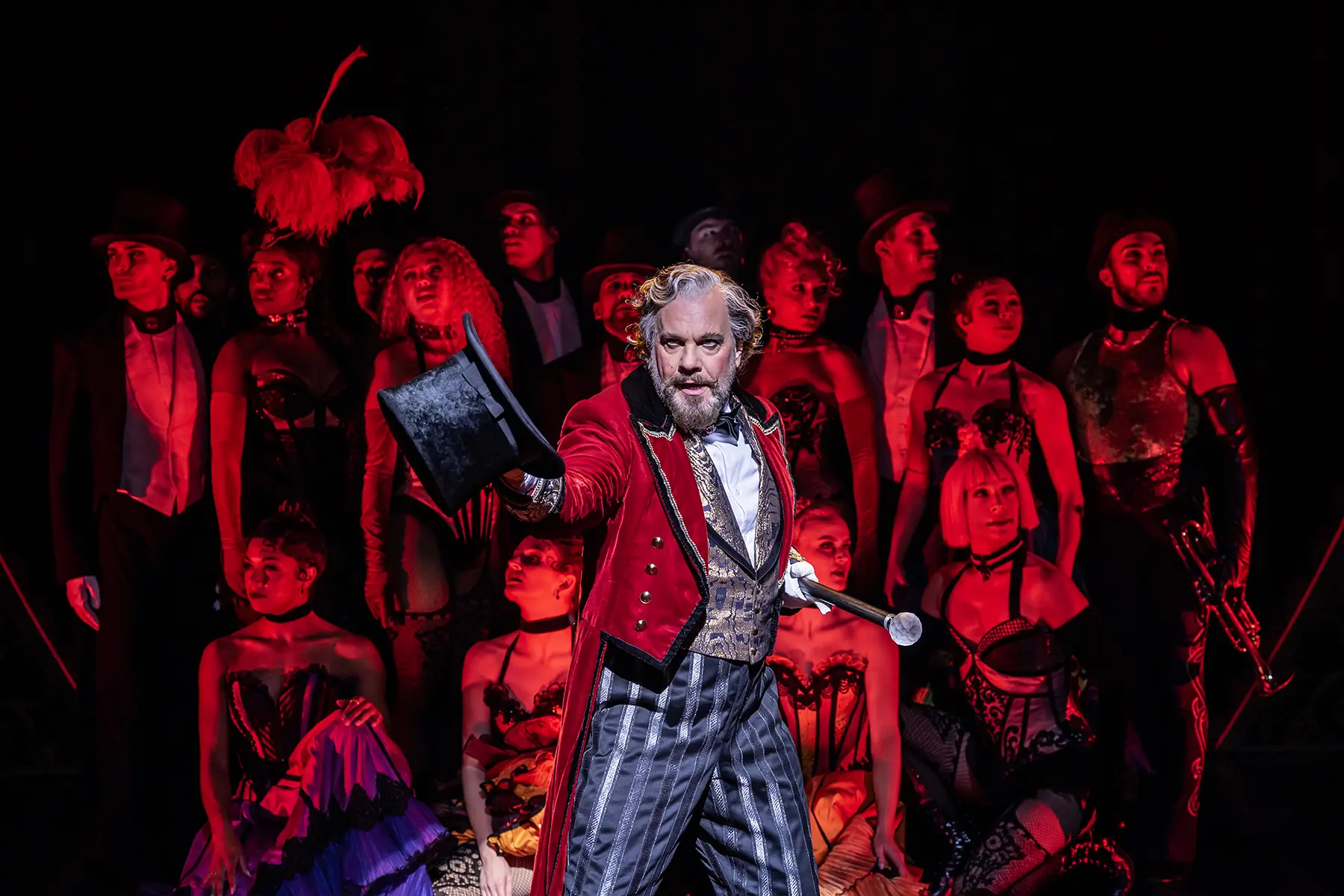 A theatrical scene from Moulin Rouge! The Musical (Netherlands production). Center stage is the character Harold Zidler, wearing a red velvet tailcoat with gold buttons, a gold-and-black patterned vest, striped pants, and holding a black top hat and cane. He is surrounded by performers in elaborate cabaret-style costumes—corsets, fishnets, top hats, and feathers—set against a dark background with dramatic red lighting.