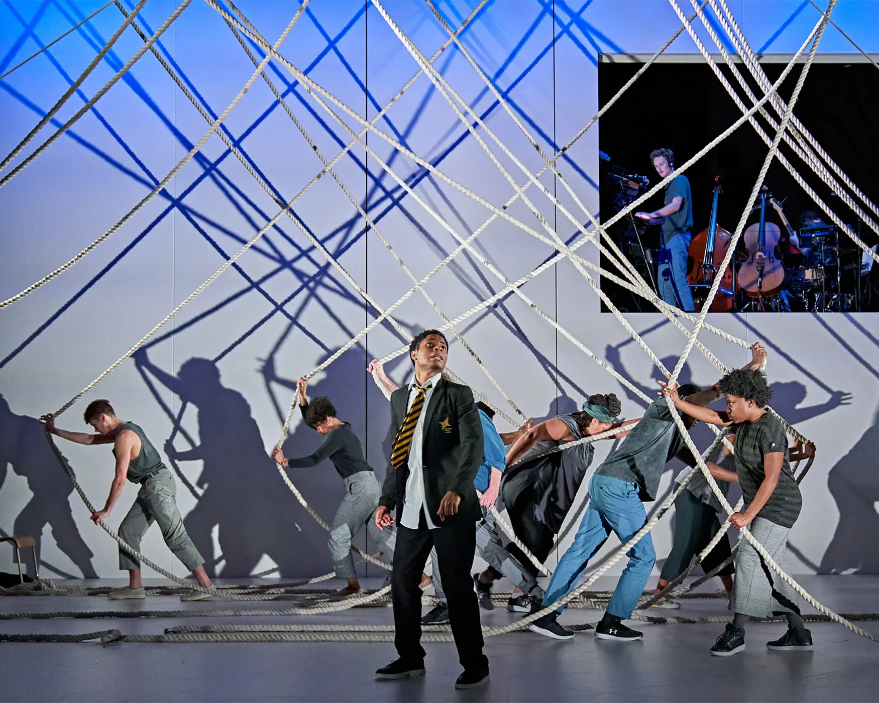 A theatrical performance featuring people interacting with a web of suspended ropes on a stage, with one person in a school uniform at the center and others in casual clothing pulling the ropes; chairs are scattered around, and a screen in the background shows musicians playing cello and double bass under dramatic lighting.