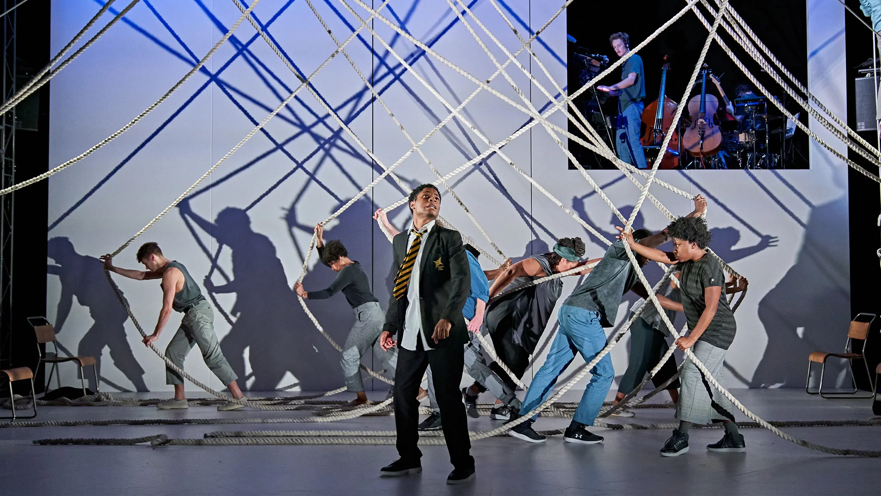 A theatrical performance featuring people interacting with a web of suspended ropes on a stage, with one person in a school uniform at the center and others in casual clothing pulling the ropes; chairs are scattered around, and a screen in the background shows musicians playing cello and double bass under dramatic lighting.