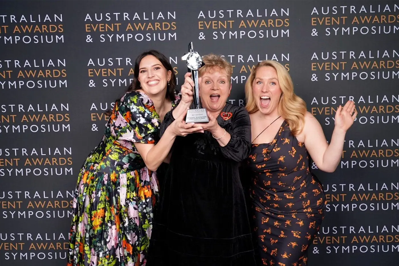 Vanessa Montalto, Dawn Martin, and Madeline Burgess pose in front of an 'Australian Event Awards' media wall, holding up a trophy.