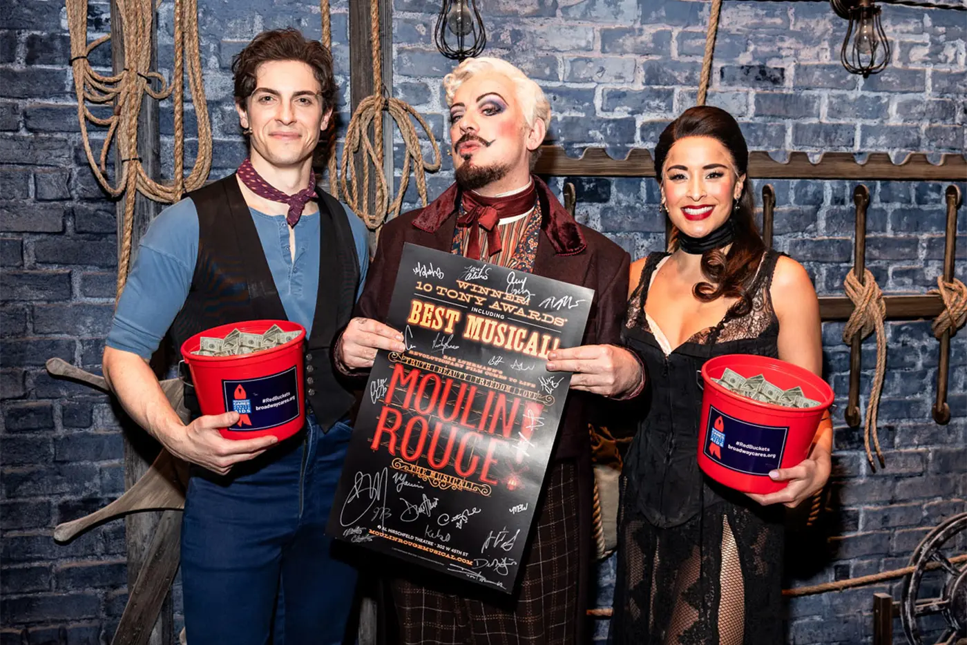 Derek Klena, Boy George, and Courtney Reed backstage holding red fundraising buckets filled with money and signed posters ready to be auctioned to raise funds for Broadway Cares / Equity FIghts AIDS.