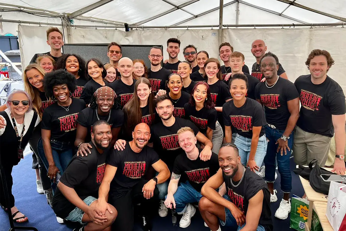 The cast of Moulin Rouge! The Musical West End at West End Live. They are smiling into the camera while wearing black-tshirts with the red Moulin Rouge! The Musical logo.