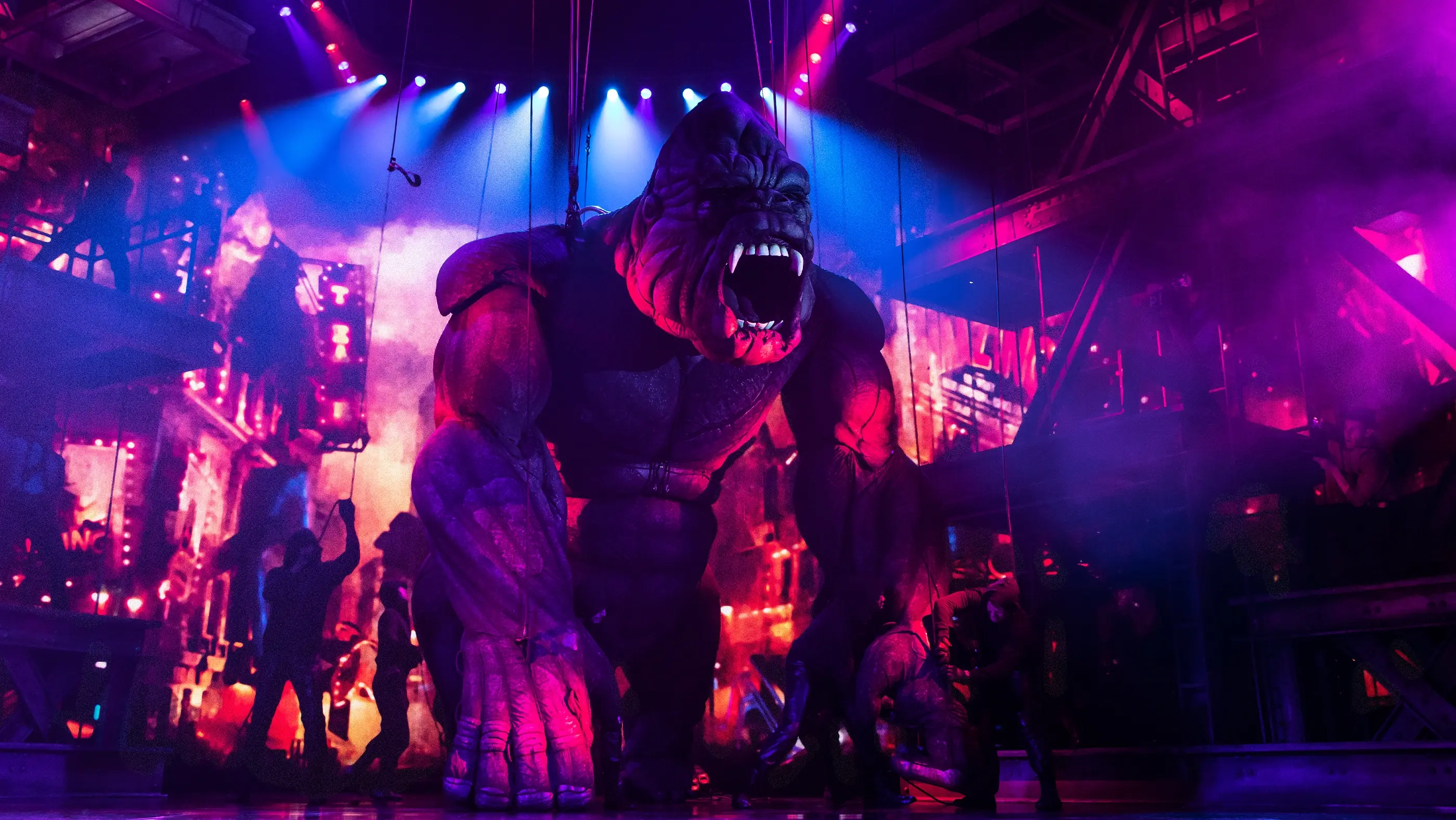 A giant gorilla puppet bathed in pink and purple light roars into the audience