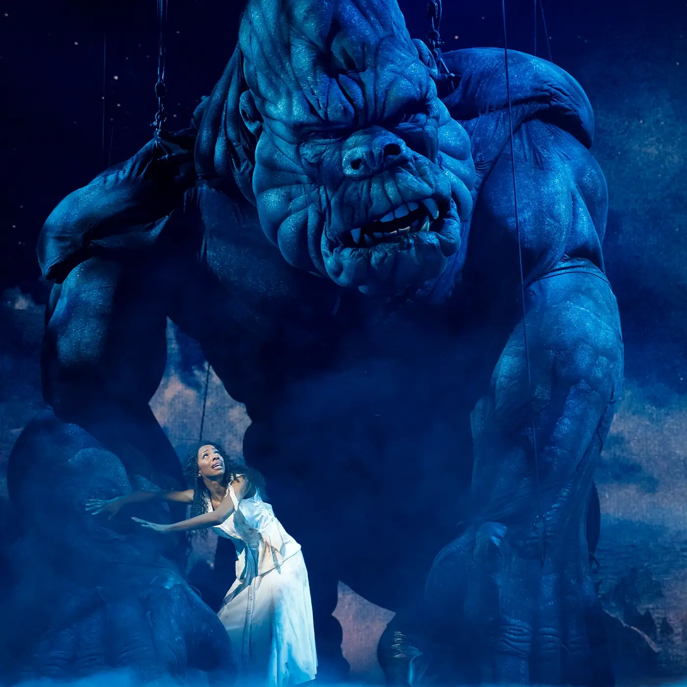 Ann Darrow stands in front of a giant King Kong on stage protecting him, blue mist light surrounds them.