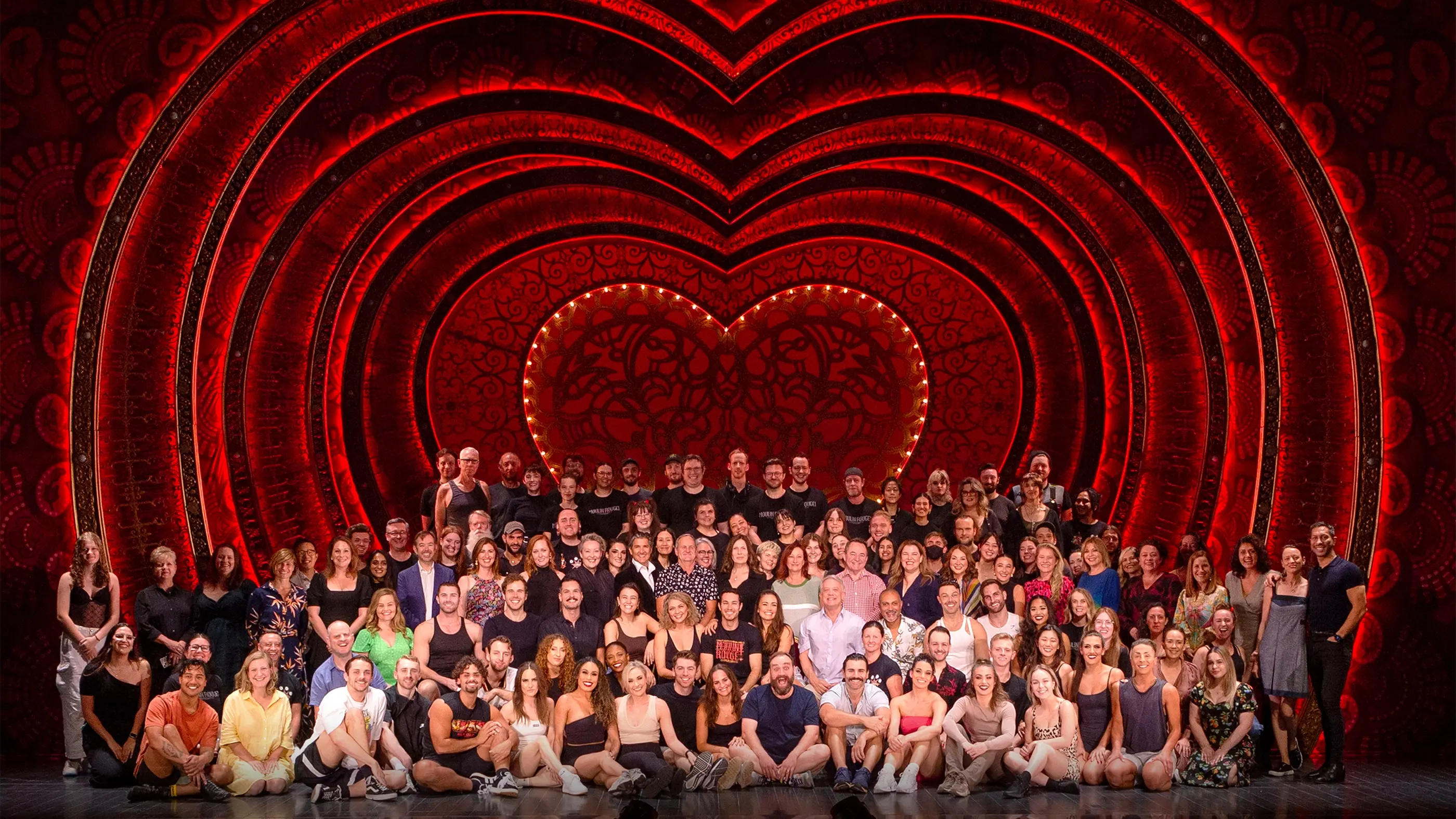 The entire company of Moulin Rouge! The Musical gathered on stage out of costume, smiling into the camera. They are surrounded by the iconic red heart portal feature of the set.