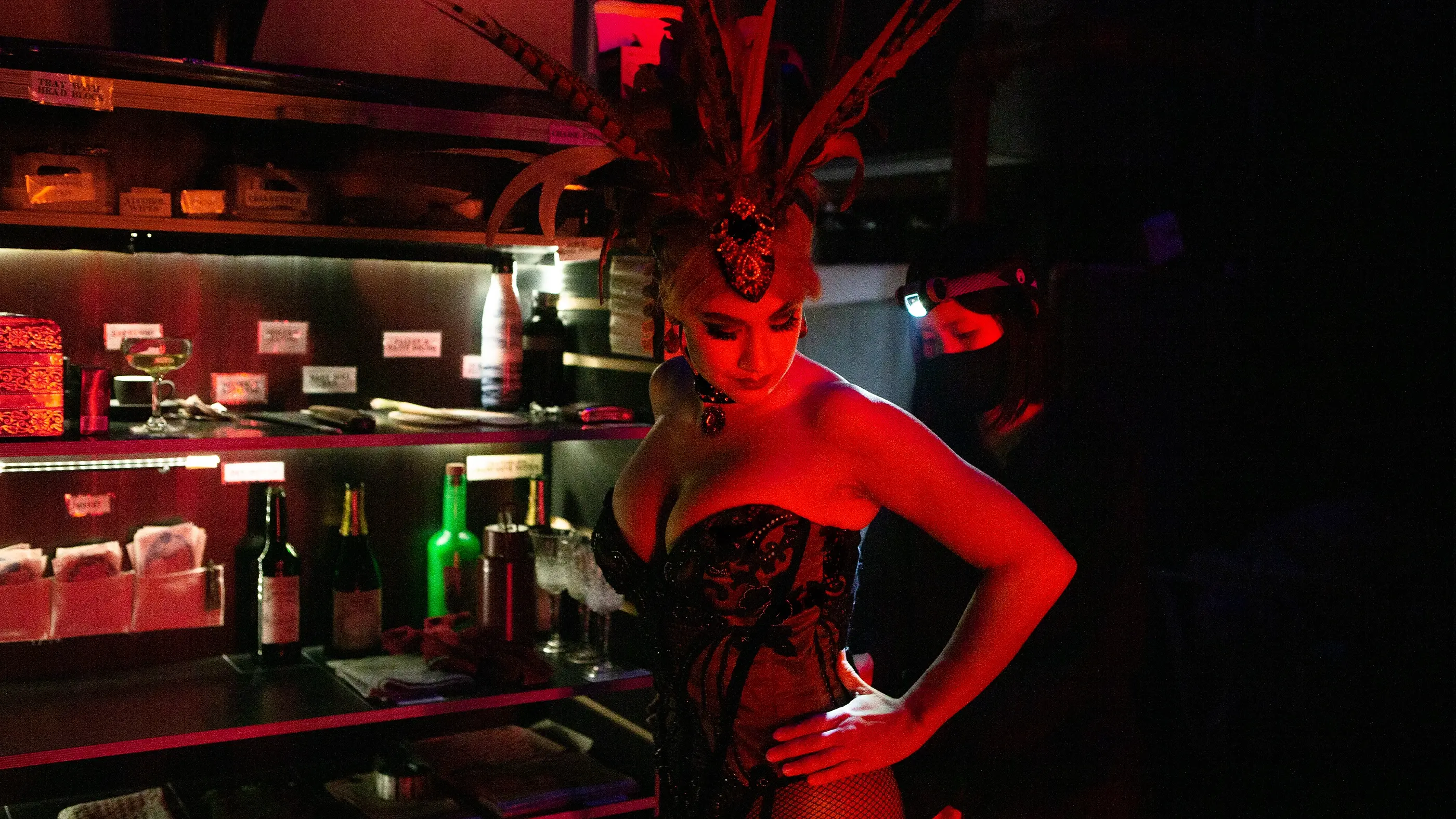 A dresser wearing a headlight assisting one of the performers in Moulin Rouge! The Musical backstage. They are lit up by red lighting, indicating the show playing off camera. Behind them is a prop stand which houses various props used throughout the production.