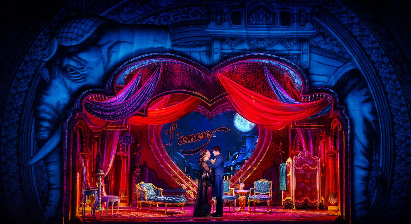Romantic stage scene from Moulin Rouge! The Musical Australia. Christian and Satine stand center stage in an intimate moment. The backdrop features a large heart, red drapery, and the word “l’amour” in cursive. Ornate furniture including chairs and a chaise lounge decorate the set. A night sky with a full moon and Parisian rooftops completes the atmospheric, love-themed setting.