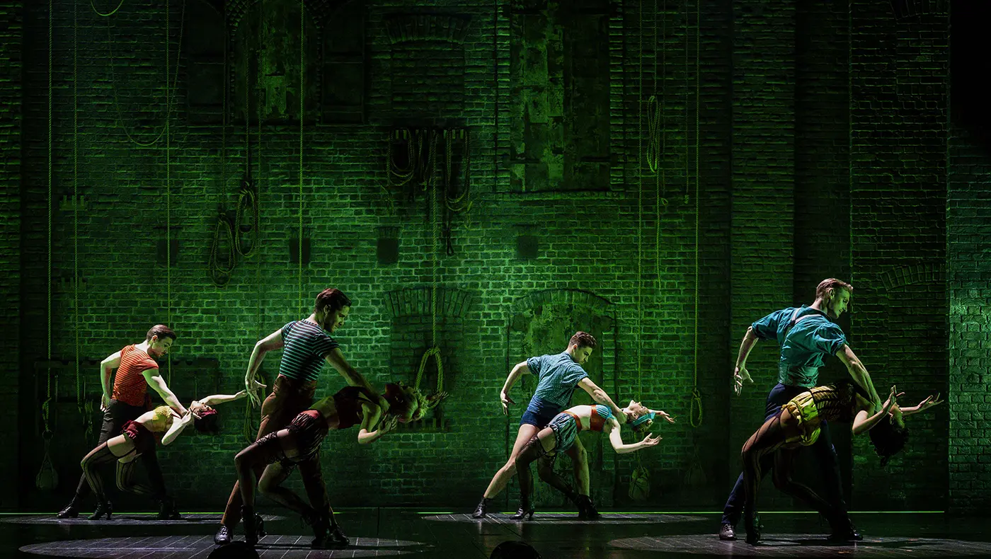 Scene from Moulin Rouge! The Musical Australia during the “Backstage Romance” number. Eight dancers perform in front of an industrial brick wall backdrop with ropes and pulleys. Green lighting casts a moody glow. Each pair strikes dramatic, synchronized poses—male dancers support their partners in deep backbends. Costumes are colorful and cabaret-inspired, including corsets, shorts, and suspenders.