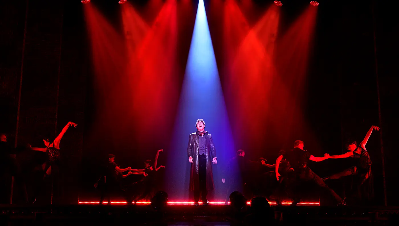 'El Tango De Roxanne' scene from from Moulin Rouge! The Musical Australia. Christian stands center stage under a bright spotlight, wearing a long coat and dark clothing. Around him, dancers strike dynamic poses, lit by red stage lights. The dark background heightens the contrast and drama of the scene.