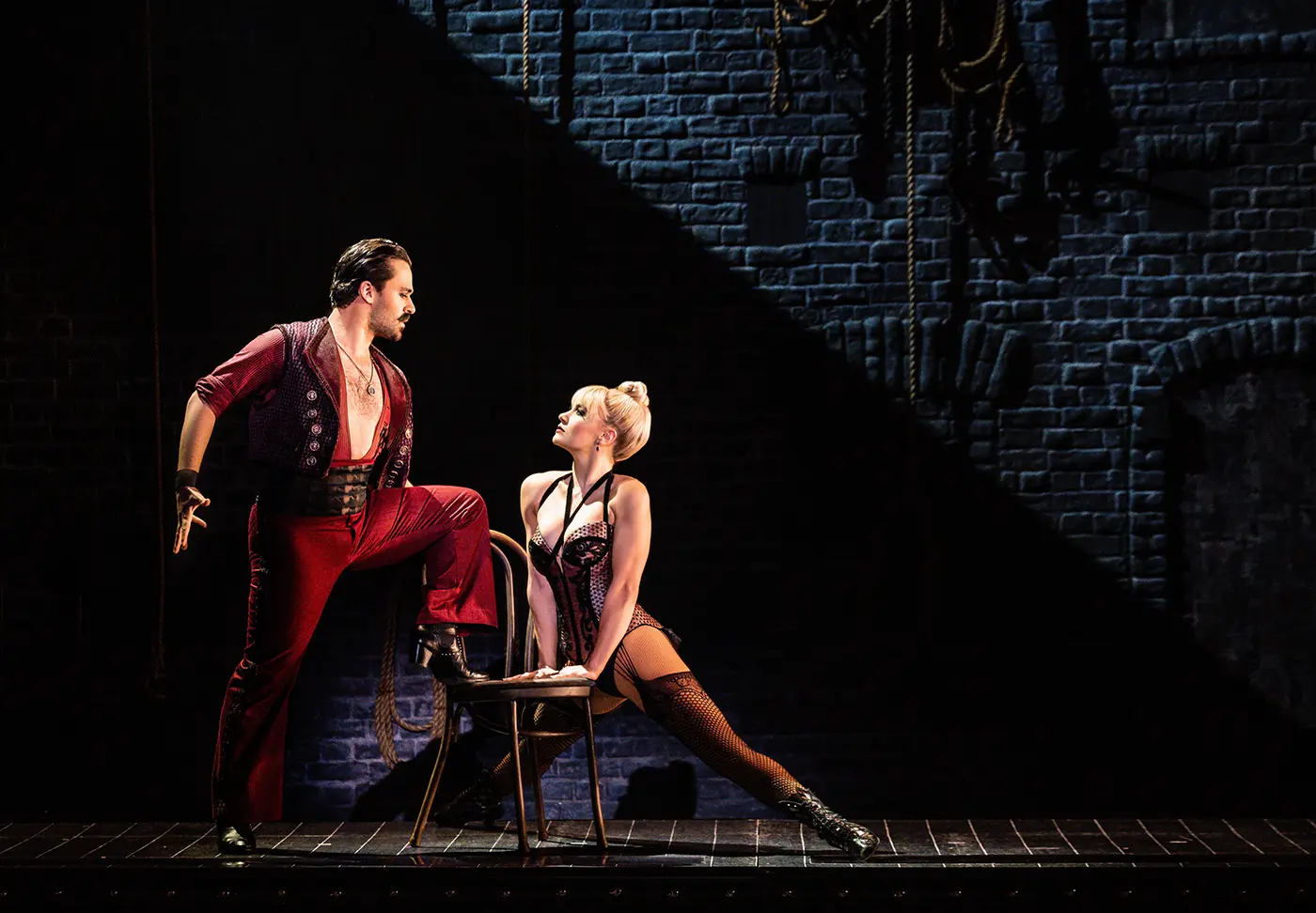 A scene from Moulin Rouge! The Musical Australia between Santiago and Nini. Santiago stands on the left with one foot on a chair, wearing a red outfit with an open shirt and black boots. Nini sits on a chair to the right, leaning forward with one leg extended, dressed in a black corseted costume with fishnet stockings and high-heeled boots. The backdrop is a brick wall with hanging ropes, evoking a backstage setting.