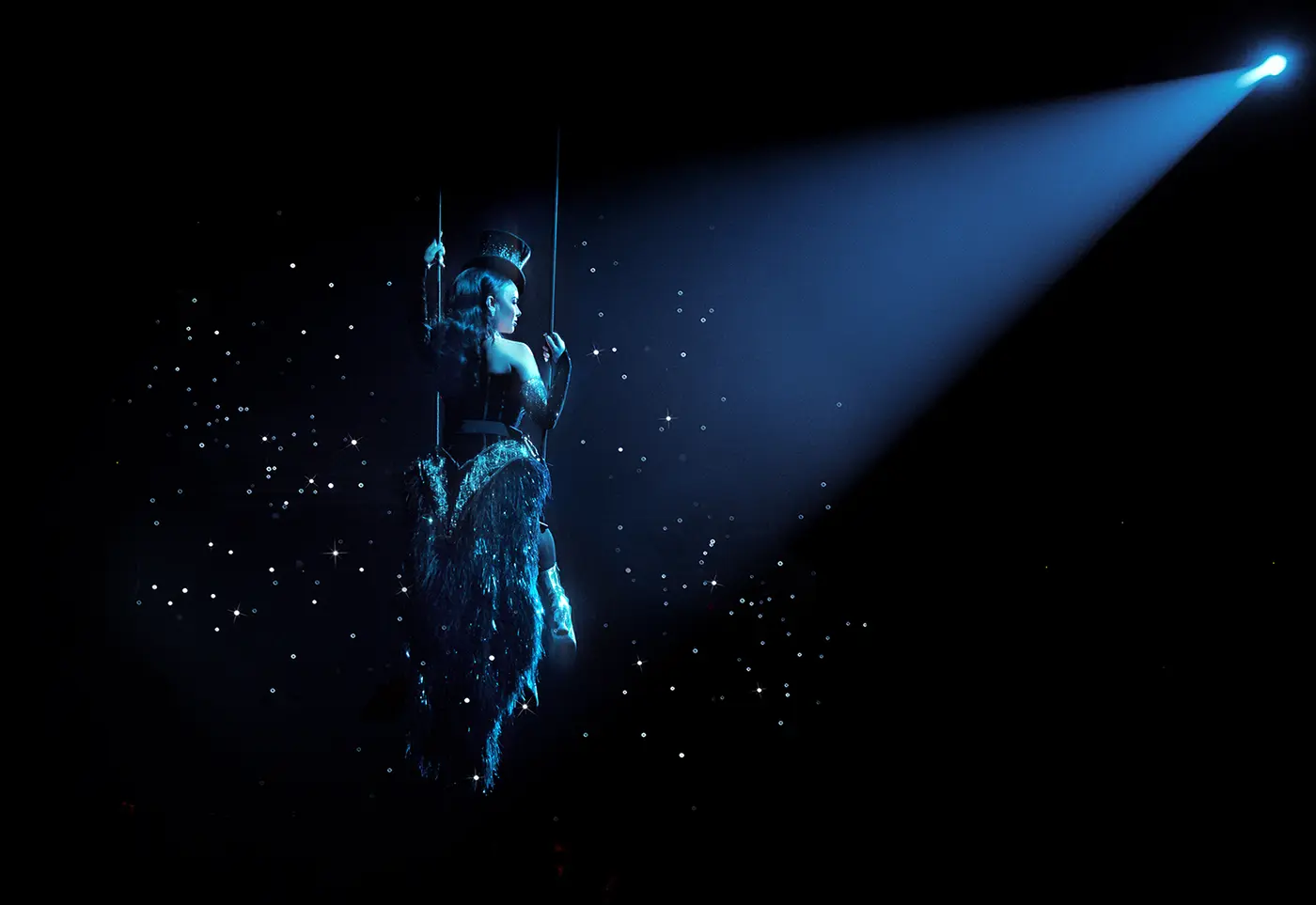 A scene from Moulin Rouge! The Musical Australia where Satine sits on a swing suspended mid-air, dressed in an elaborate corseted costume. A spotlight from the right highlights the performer against a dark backdrop filled with scattered lights resembling stars, creating a dramatic and ethereal atmosphere.