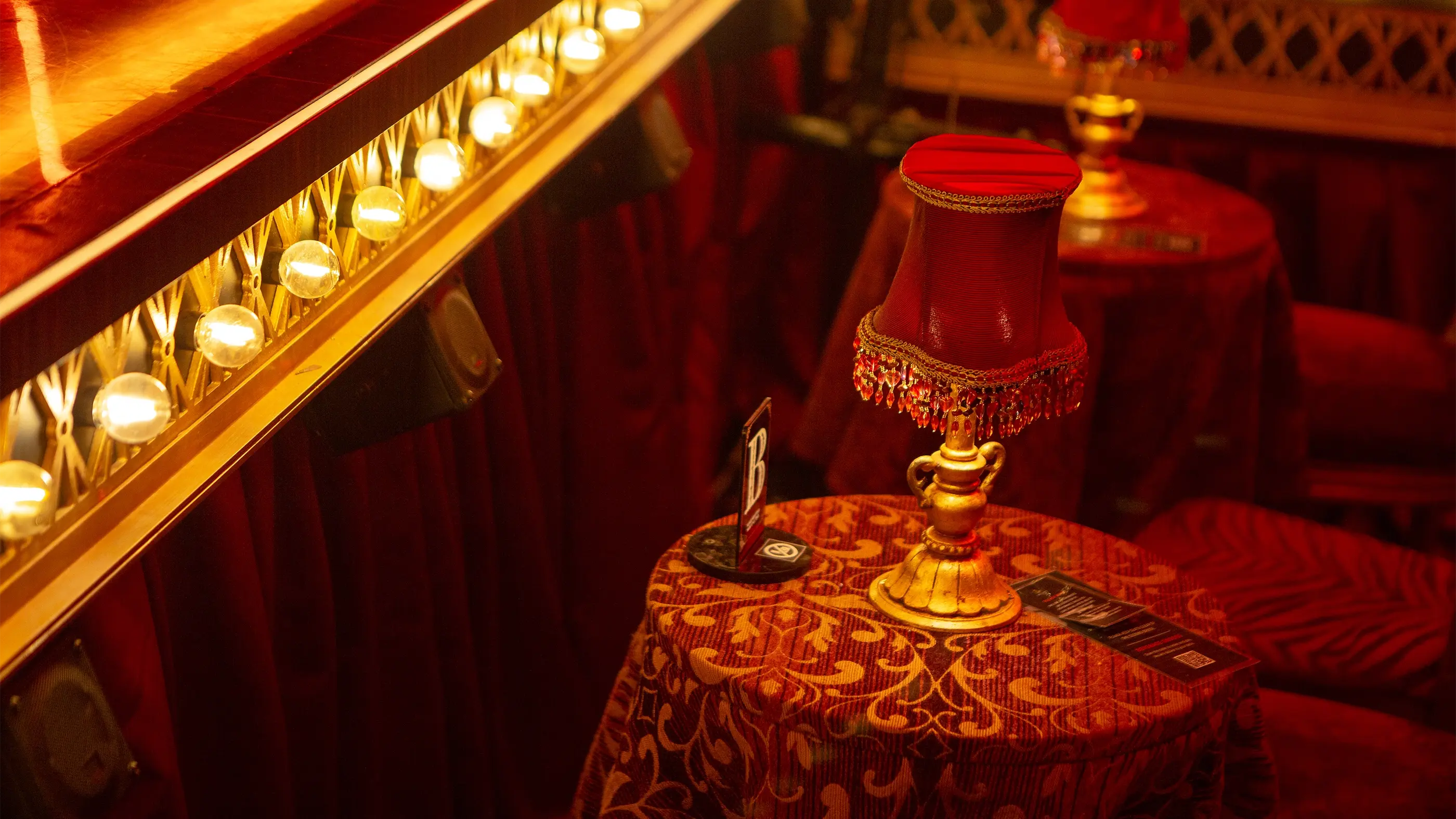 A table in the special Can-Can seating area of Moulin Rouge! The Musical. The table is draped in a red and gold patterned cloth and holds a small red-shaded lamp with a gold base, a menu or program, and a sign marked with the letter “B.” Surrounding the table are plush red seats and decorative lighting along the stage edge.