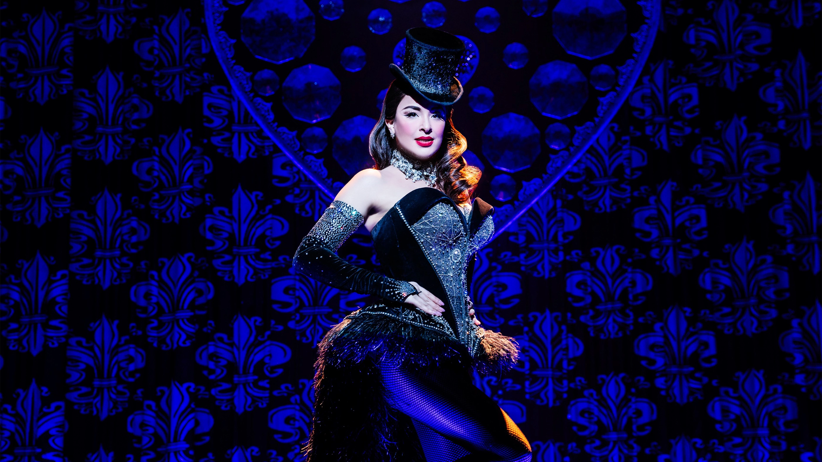 Courtney Reed as Satine in Moulin Rouge! The Musical performing 'The Sparkling Diamond' in a sparkling corseted dress and top hat, surrounded by a blue-lit set.