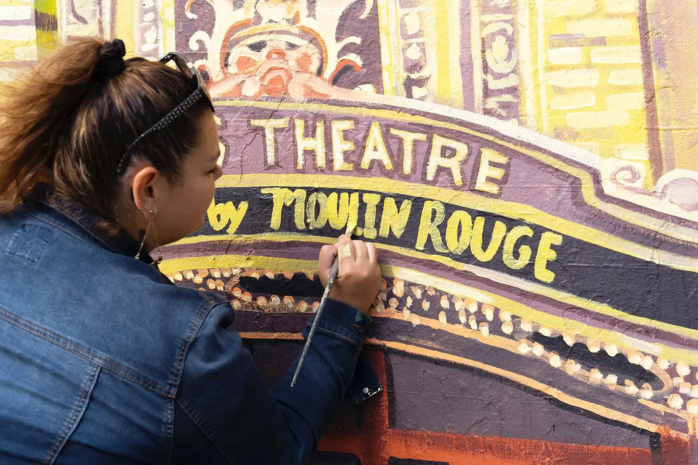 A person applying the finishing touches to a mural of a theatre showing Moulin Rouge! The Musical.