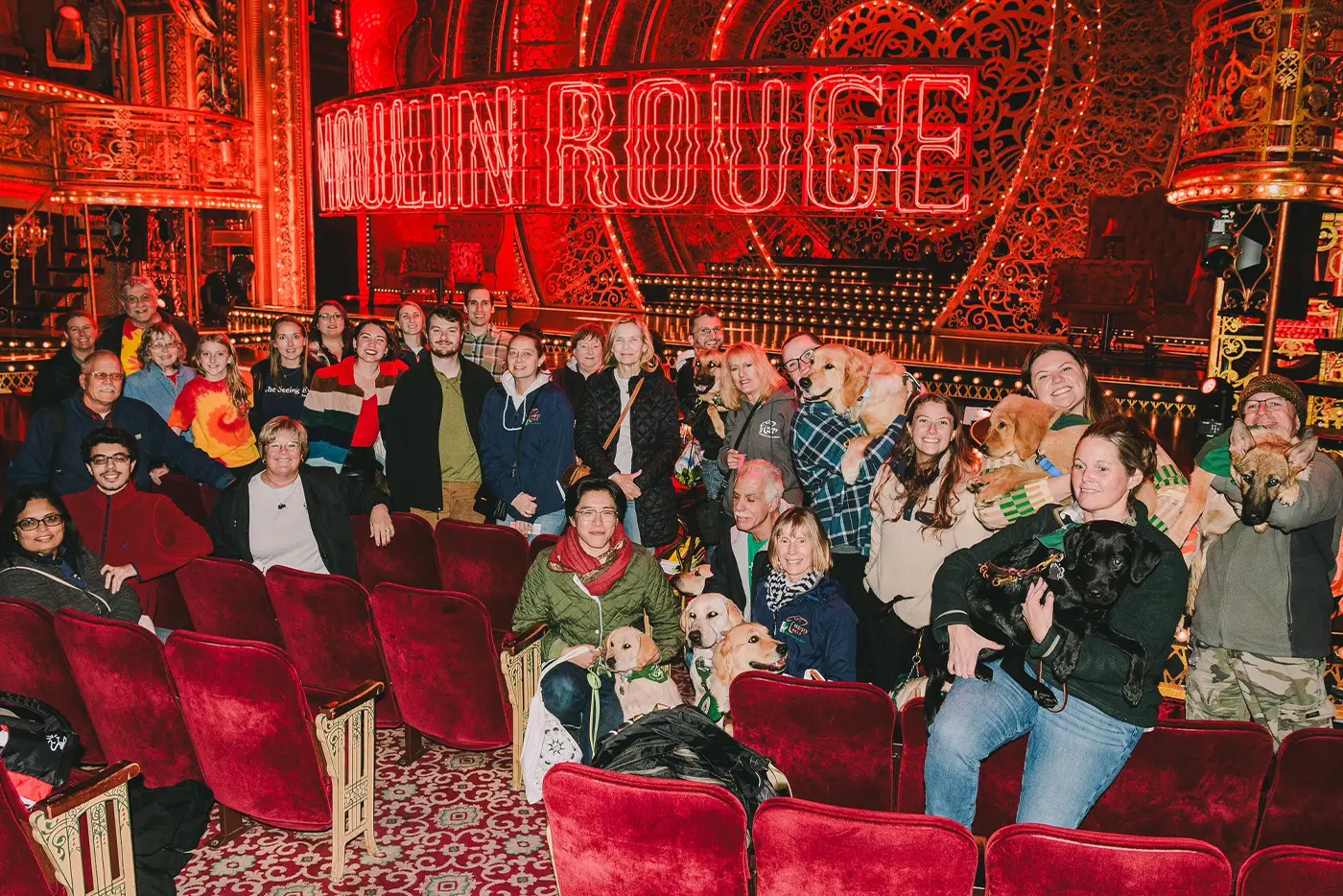 A group of audience members posing from their seats in front of the Moulin Rouge! The Musical set, holding their Guide Dogs of various breeds.