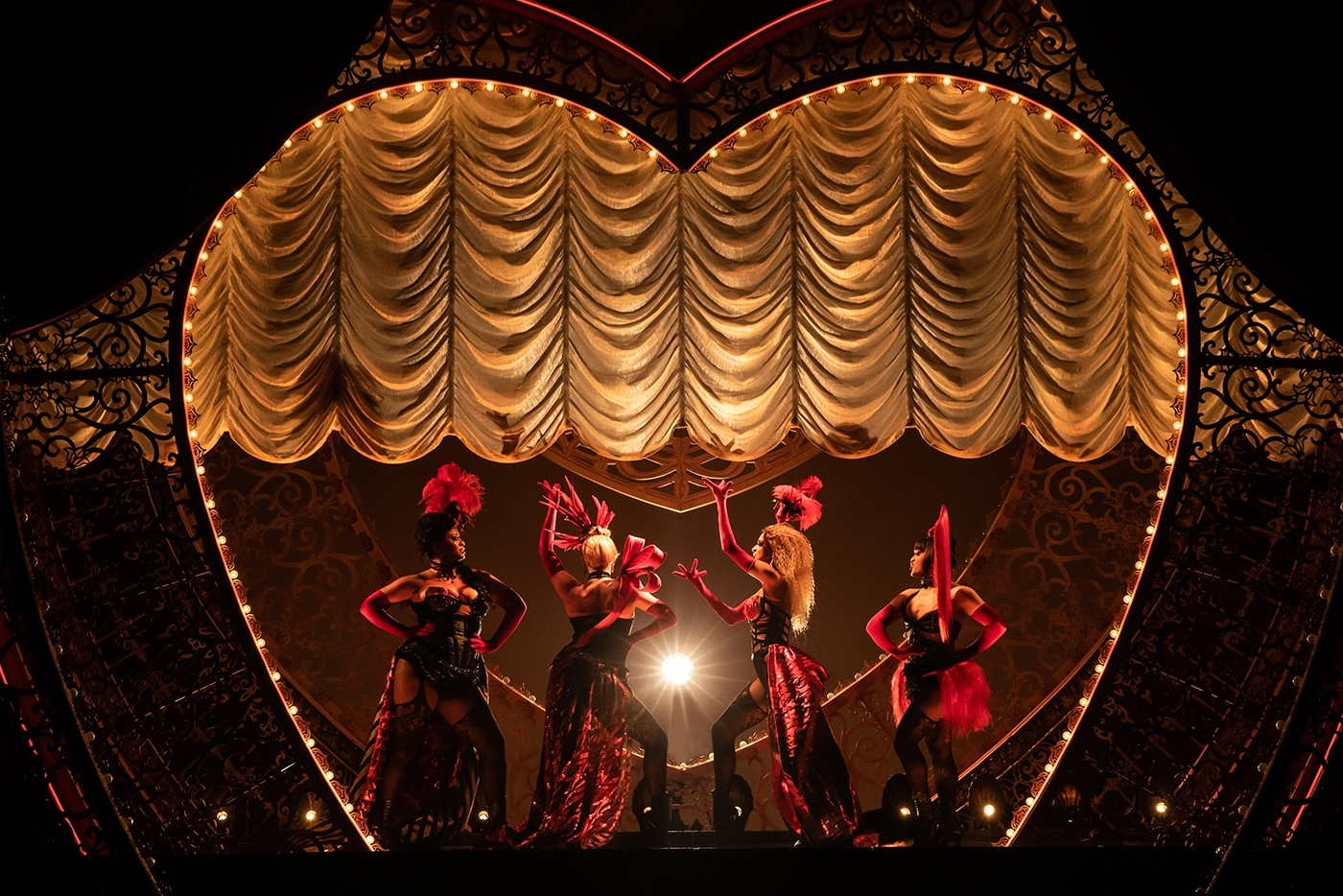 Scene from Moulin Rouge! The Musical on Broadway featuring the Lady M’s in red and black burlesque costumes with feathered headpieces, posed in front of an ornate heart-shaped stage backdrop.