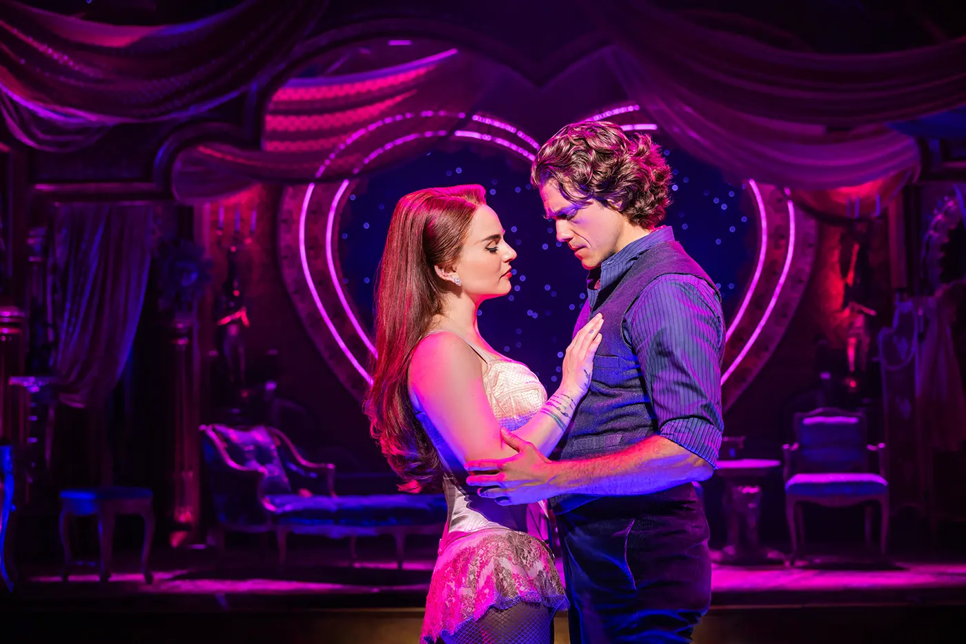 Scene from the Moulin Rouge North American tour in Satine’s dressing room, with two characters, Christian and Satine, played by Aaron Tveit and Jojo, embracing while surrounded by opulent furnishings and purple drapes.