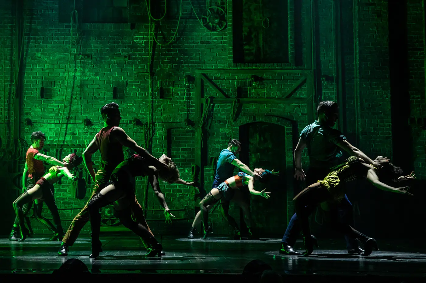 Scene from Moulin Rouge! The Musical during the “Backstage Romance” number. Eight dancers perform in front of an industrial brick wall backdrop with ropes and pulleys. Green lighting casts a moody glow. Each pair strikes dramatic, synchronized poses—male dancers support their partners in deep backbends. Costumes are colorful and cabaret-inspired, including corsets, shorts, and suspenders.