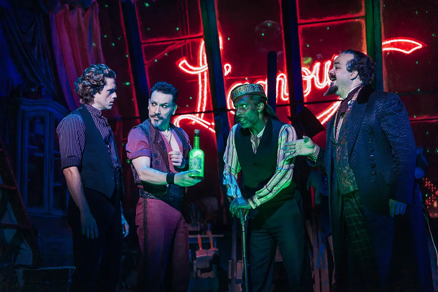 'Chandelier' scene from Moulin Rouge! The Musical. Christian, Santiago, Toulouse-Lautrec, and Harold Zidler stand together under green lighting. Santiago holds a green, glowing bottle of Absinthe. Behind them, draped fabric and a large neon sign reading “L’amour” in red cursive letters create a whimsical and moody backdrop.