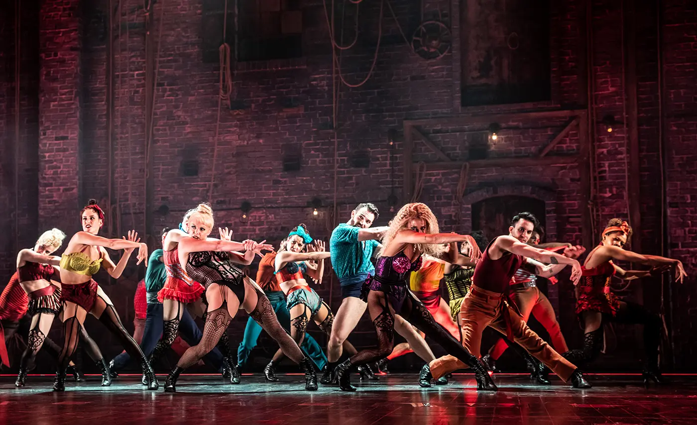 'Backstage Romance' scene from Moulin Rouge! The Musical (Broadway cast) featuring ensemble cast are dancing in sync in front of an industrial brick wall backdrop with ropes and pulleys. Costumes are colorful and cabaret-inspired, including corsets, shorts, and suspenders.