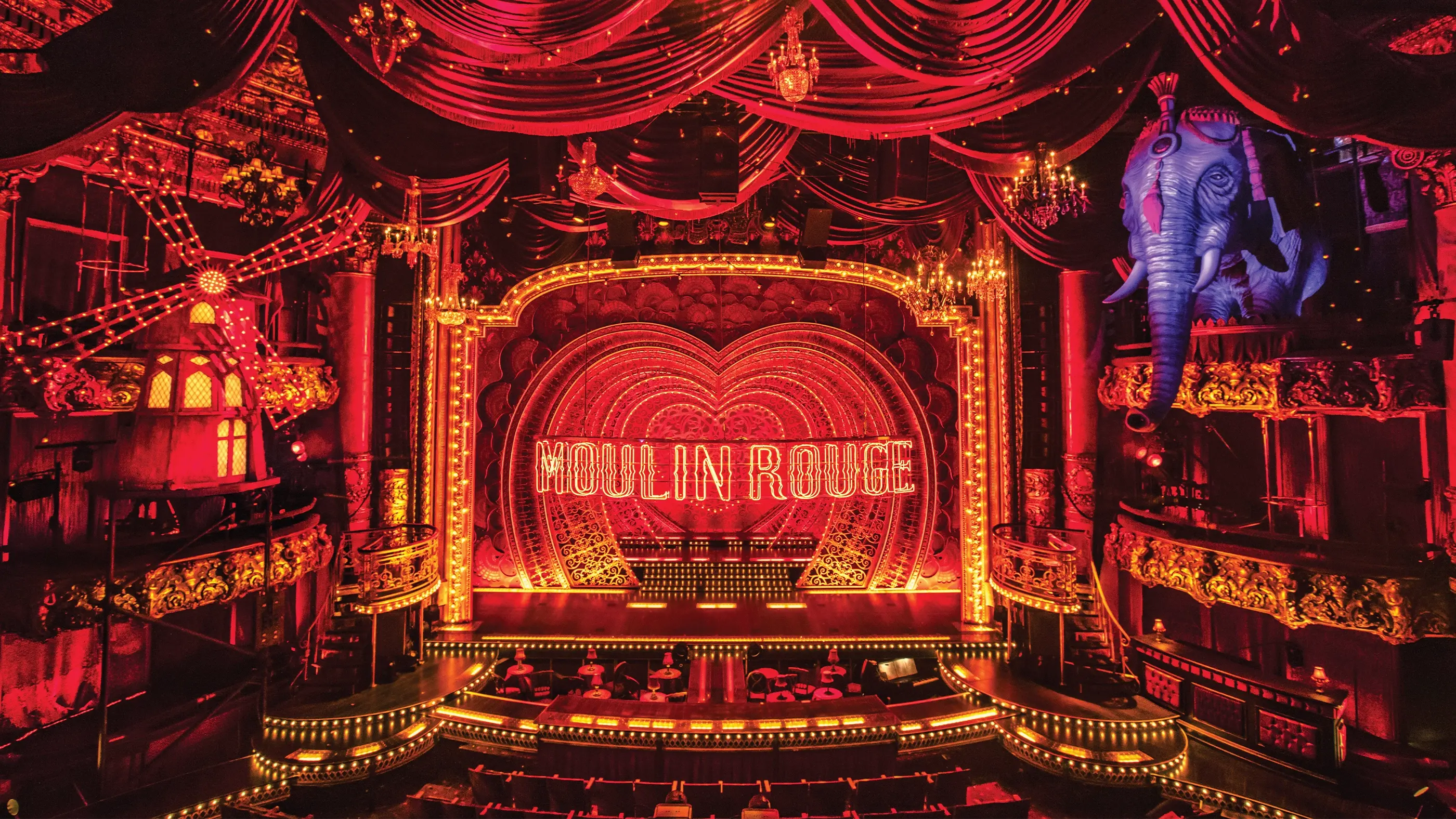 A stage set from Moulin Rouge! The Musical, featuring a richly decorated interior with red velvet drapes, ornate chandeliers, and a glowing 