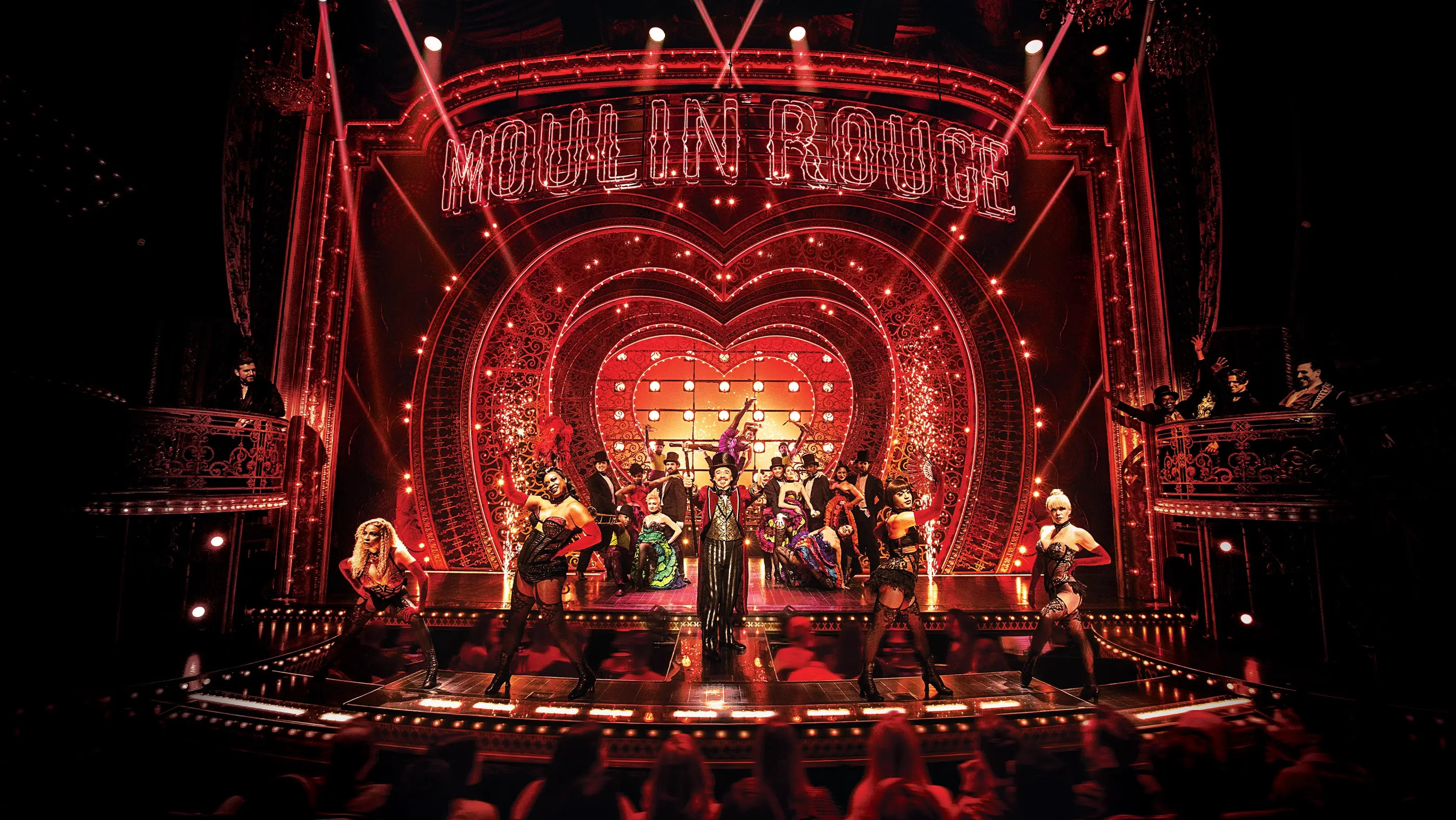 Scene from Moulin Rouge! The Musical on Broadway, featuring dancers in extravagant costumes on a lavish red and gold stage with a heart-shaped illuminated backdrop and “MOULIN ROUGE” marquee.