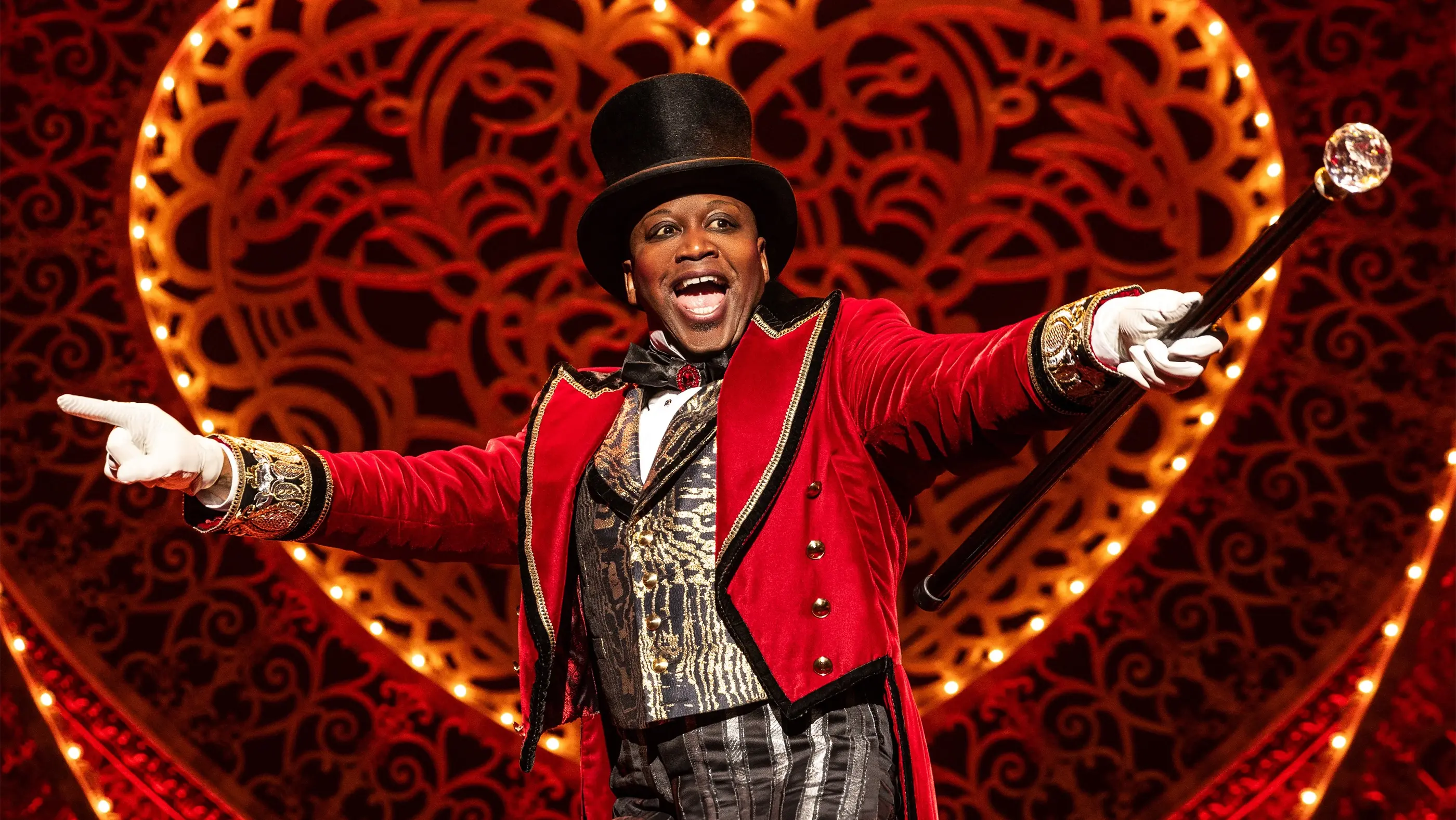 Titus Burgess as Harold Zidler from Moulin Rouge! The Musical on Broadway. He wears a red and gold ornate jacket, black and white striped pants, a top hat, and holds a cane.