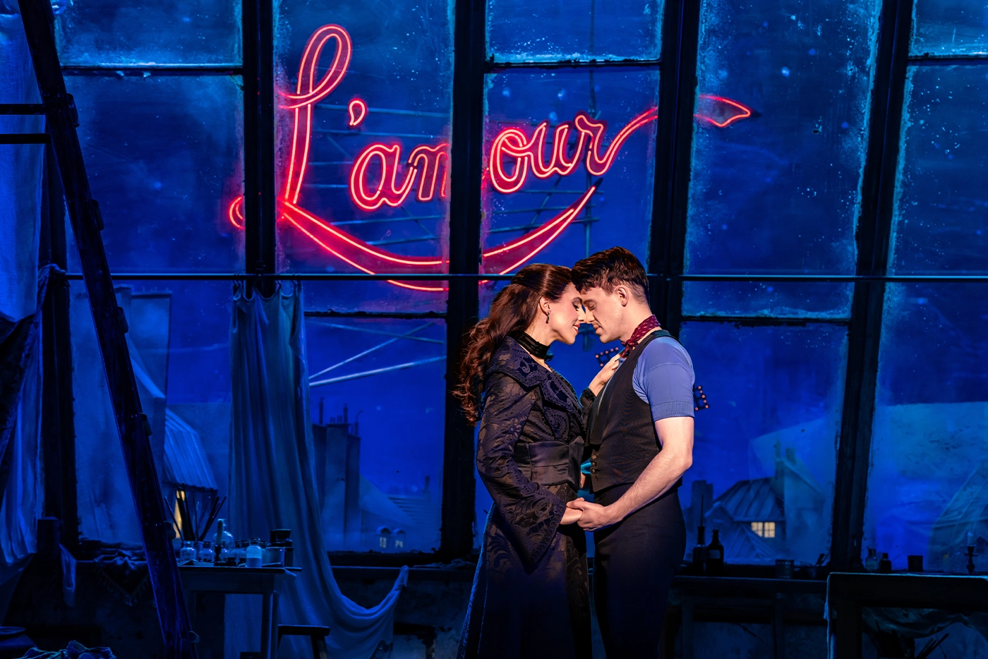 Scene from Moulin Rouge! The Musical featuring the German cast in “Come What May,” with two characters holding hands under blue lighting, framed by large windows and a glowing “L’amour” sign.