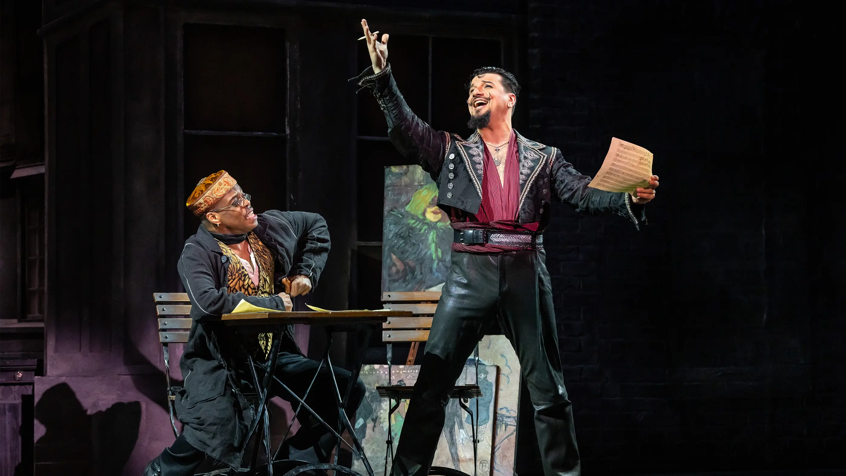 Scene from Moulin Rouge! The Musical (Germany) featuring Toulouse-Lautrec and Santiago. Toulouse-Lautrec, seated on the left, wears a dark coat and hat and sits at a table with papers. Santiago stands on the right, holding sheet music in one hand and gesturing with the other. He wears black pants, a red shirt, and a detailed jacket. The background includes dark walls and an easel with artwork.