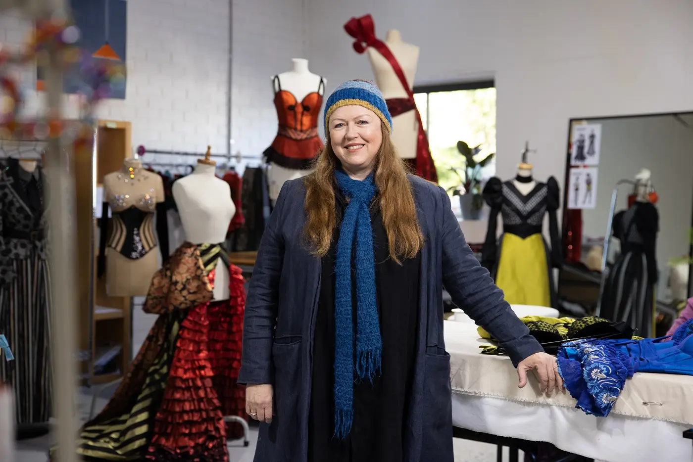 Janet Hine, Costume Design Associate of Moulin Rouge! The Musical Australia, smiling into the camera. She is surrounded by various costumes for the show on mannequins.