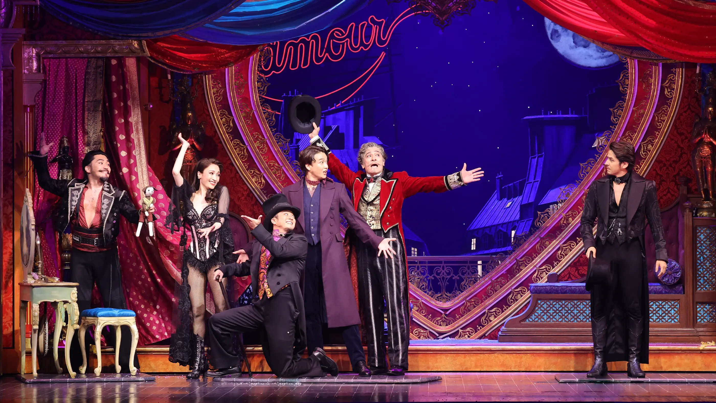 Scene from Moulin Rouge! The Musical (Japan), during the “Pitch Song.” The stage features red and blue drapes, ornate golden frames, and a backdrop of a moonlit Parisian rooftop. A neon “L'amour” sign glows at the top center. Six performers in are on stage: one on the far left holds a doll, another in black lace strikes a pose, two center-stage performers interact with a hat, and two on the right wear formal attire.