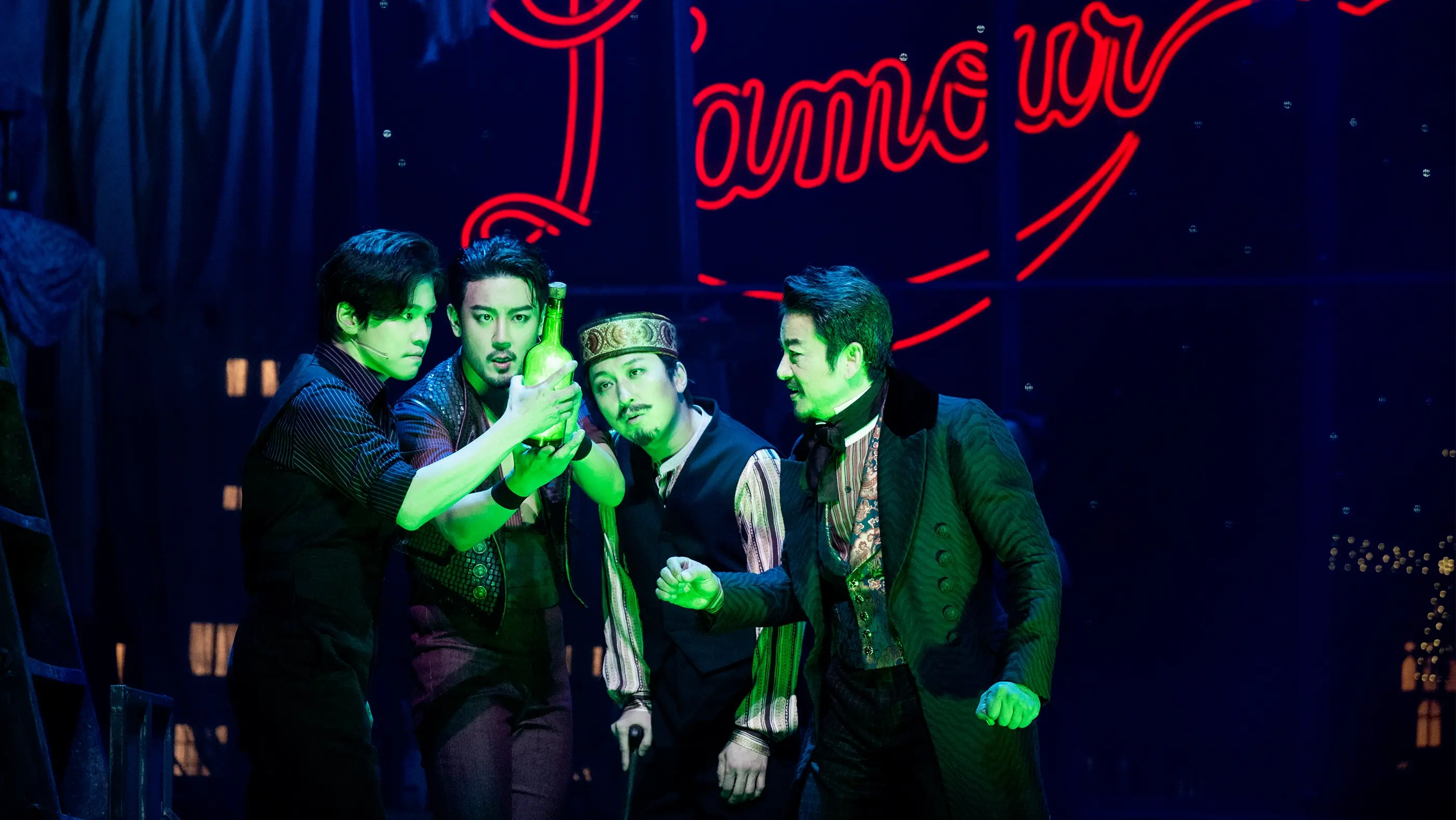 'Chandelier' scene from Moulin Rouge! The Musical (Korea). Four performers stand together under green lighting, appearing to hold or examine a bottle of Absinthe. Behind them, draped fabric and a large neon sign reading “L’amour” in red cursive letters create a romantic and moody backdrop.