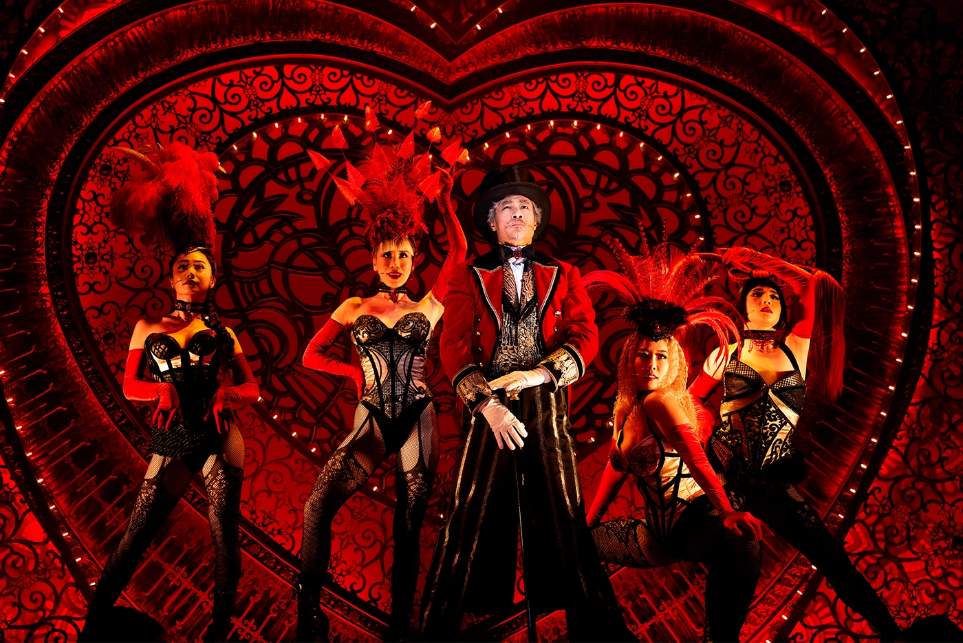 Scene from Moulin Rouge! The Musical Korea featuring Zidler in a red and black costume with top hat, surrounded by Lady M’s in feathered showgirl attire against a heart-shaped, red-lit backdrop.