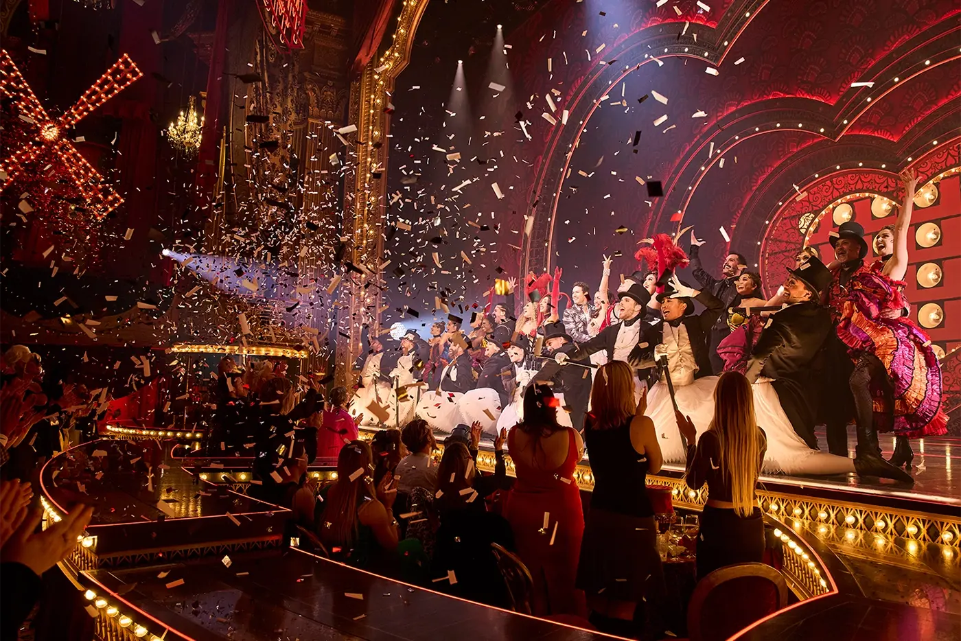 Curtain call scene from Moulin Rouge! The Musical. Performers line up on a lavishly decorated stage. Confetti falls from above as the cast takes their bows. The audience is visible in the foreground, applauding the performance.