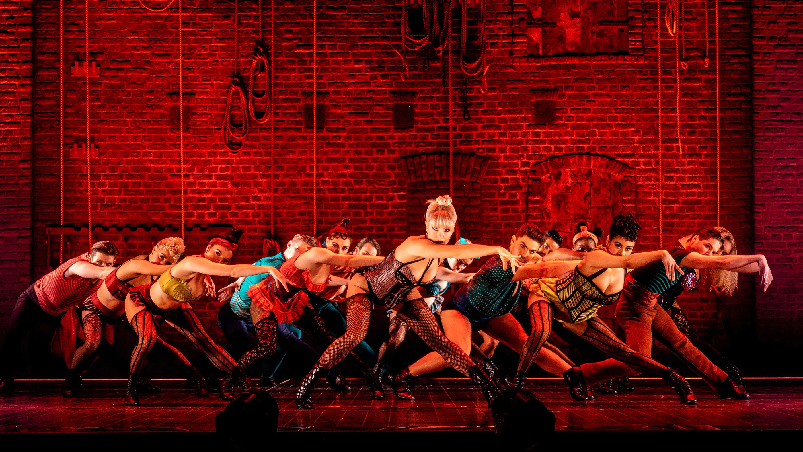 Scene from Moulin Rouge! The Musical (Australia) during “Backstage Romance.” A group of dancers performs in front of a red brick wall with ropes hanging down, evoking a backstage atmosphere. The dancers wear colorful, cabaret-style costumes and strike dynamic poses with arms extended forward, capturing the energy of the number.