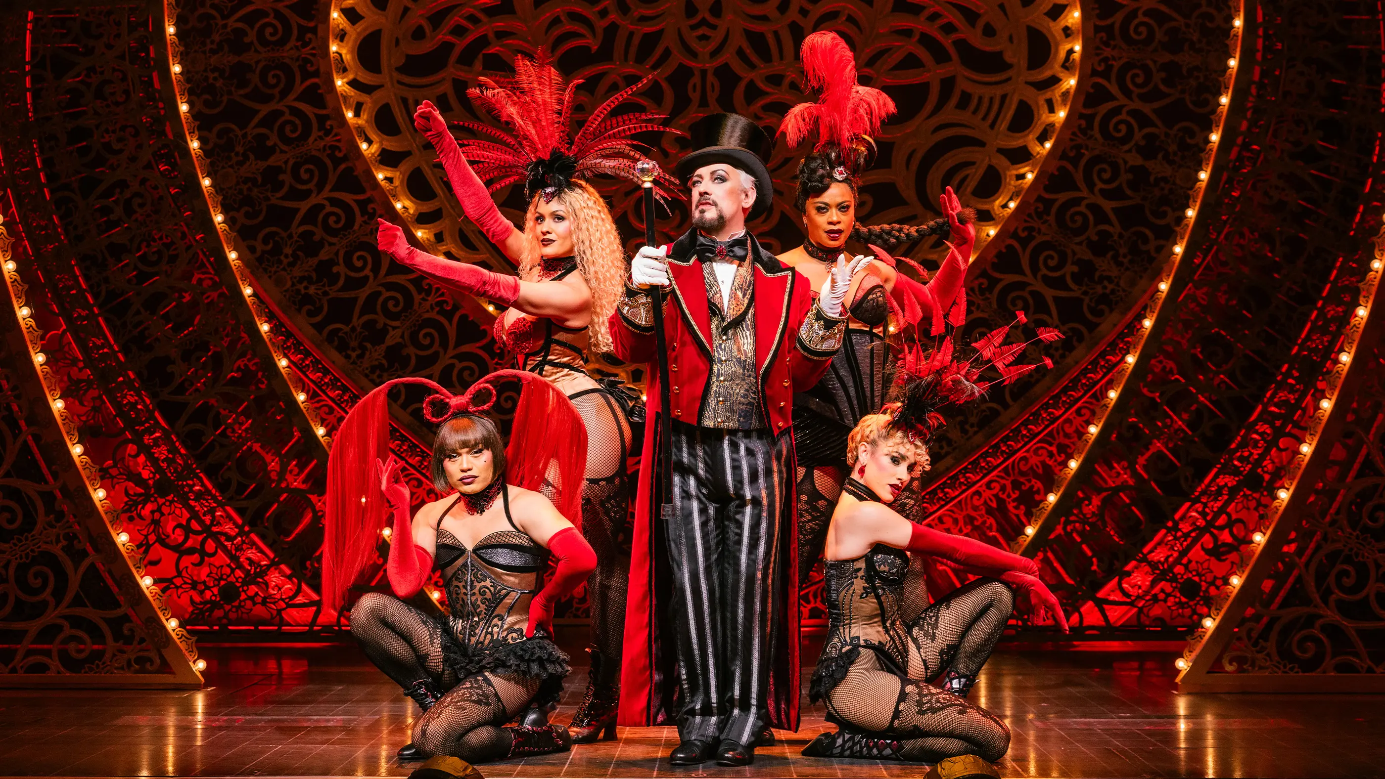 Scene from Moulin Rouge! The Musical (Broadway) featuring Boy George as Harold Zidler center stage. He wears a red and gold ornate jacket, black and white striped pants, a top hat, and holds a cane. Surrounding him are the four Lady Ms, dressed in corsets, fishnet stockings, gloves, and feathered headpieces. The background is richly decorated with red lighting and intricate patterns.