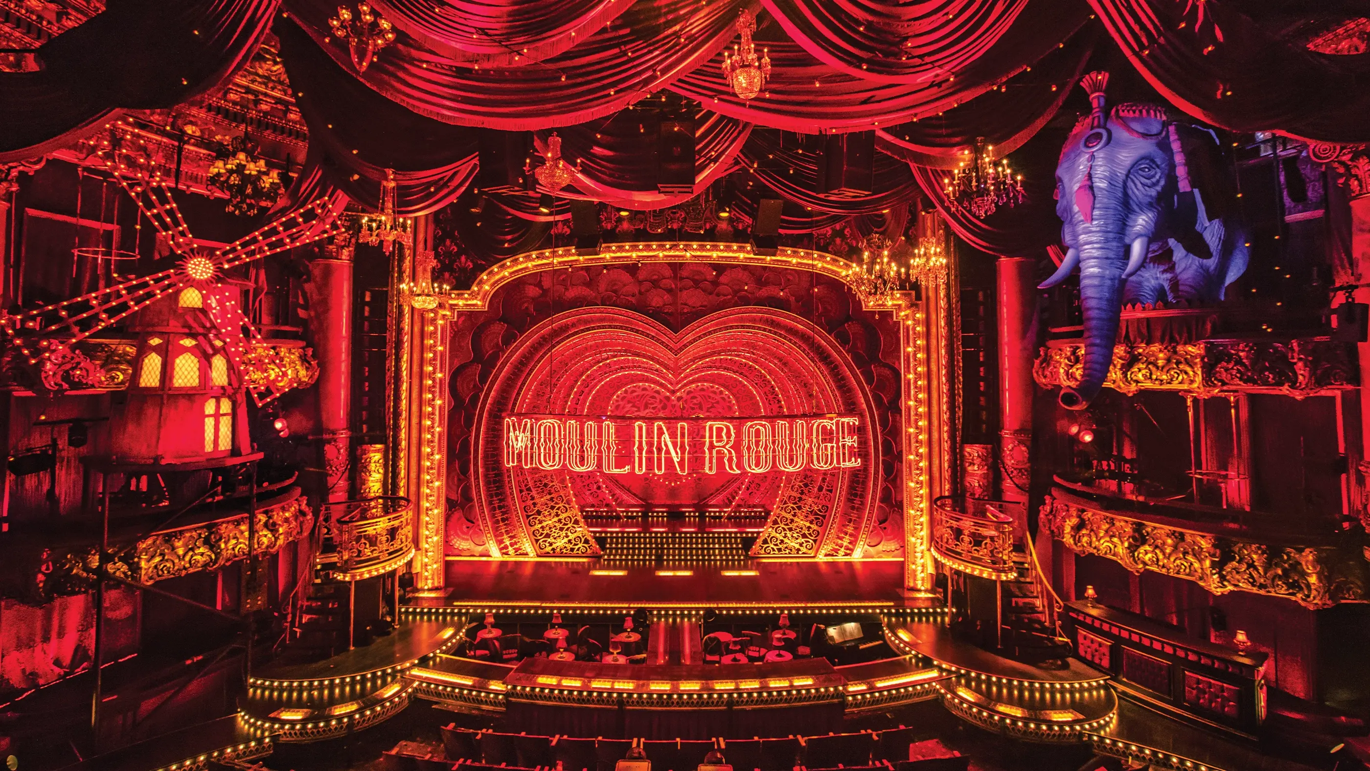 A stage set from Moulin Rouge! The Musical, featuring a richly decorated interior with red velvet drapes, ornate chandeliers, and a glowing 
