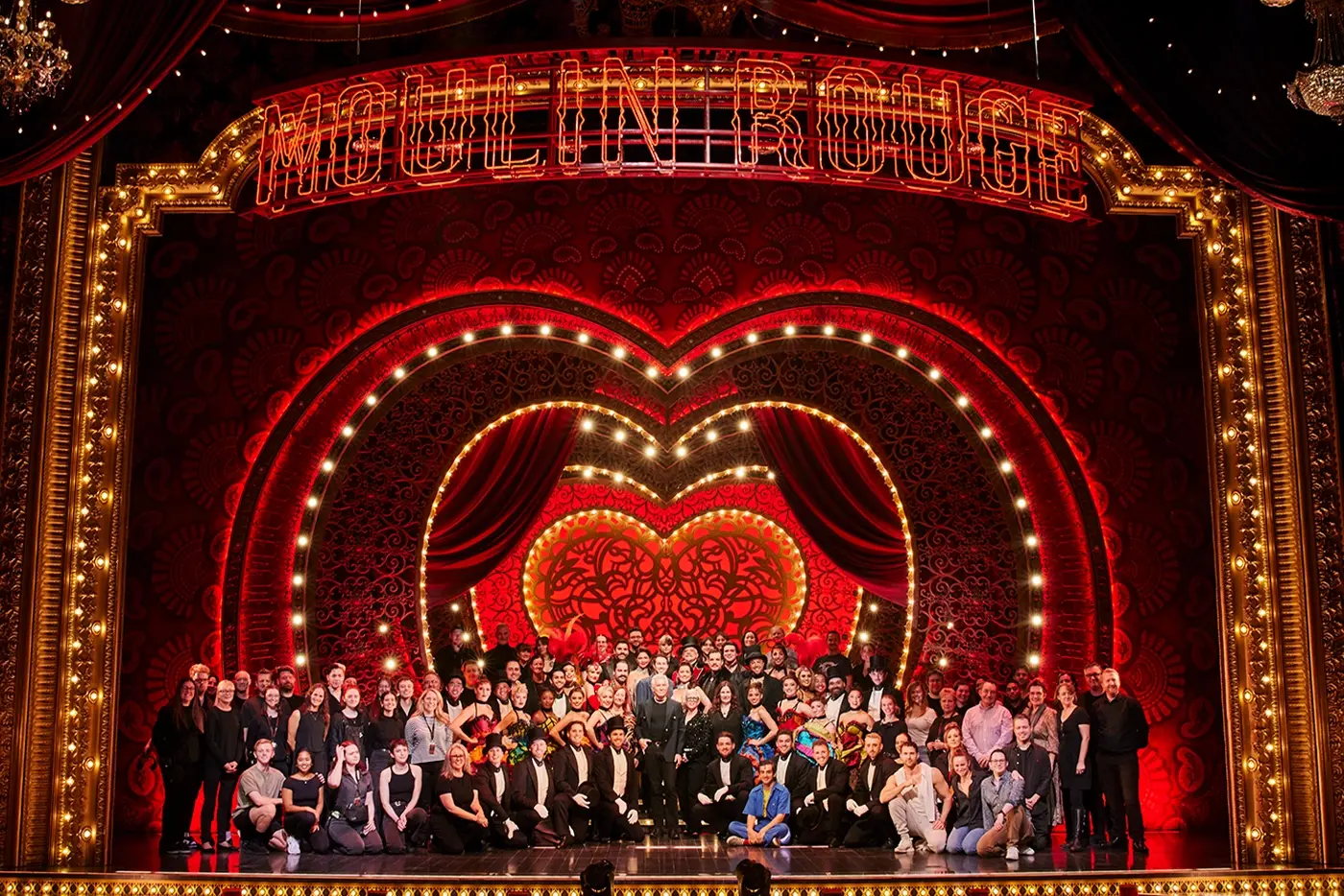 Cast and crew of Moulin Rouge! The Musical are grouped together on a lavishly decorated stage featuring chandeliers, red drapes, golden accents, and a large illuminated “MOULIN ROUGE” sign. Baz Luhrmann, Catherine Martin, and Carmen Pavlovic are pictured in the center of everyone.